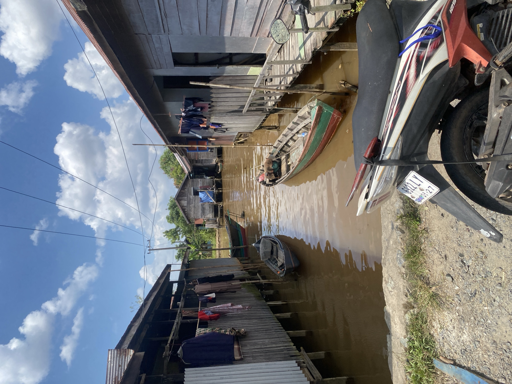 The image depicts a waterfront scene in Berangas Timur, Indonesia, likely a village on the coast or a river. Two rows of wooden stilted houses line a narrow waterway. Laundry hangs on lines strung between houses on the left. Several small wooden boats are moored in the brown water, some close to the houses and others further out. One larger boat with red and green trim is prominent in the middle ground. The sky is blue with scattered white clouds. A portion of a motorcycle, with a license plate visible that reads "6922 LY 08-27", is in the foreground. The overall impression is of a traditional, water-based community.