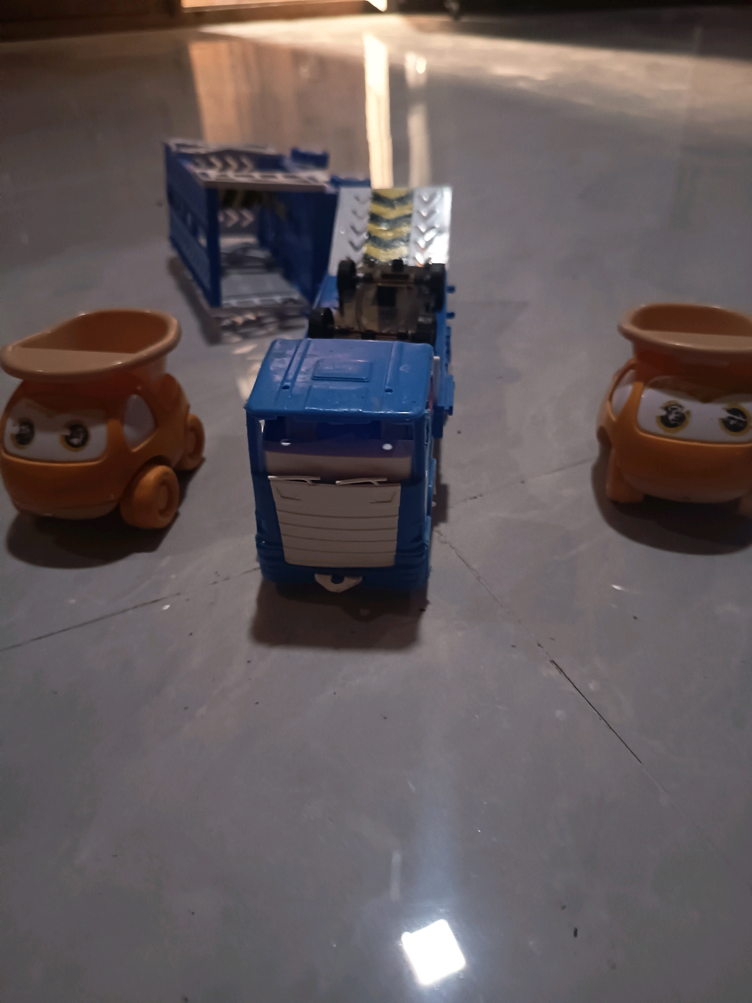 In Sidorejo, Indonesia, a toy car transporter is positioned on a tiled floor, with two small orange toy cars on either side. The transporter, predominantly blue, has a ramp at the rear, decorated with yellow and black hazard stripes. The orange toy cars, featuring stylized facial expressions with large eyes, face the transporter. The floor appears to be a neutral gray tile, with some visible seams and a reflection of light in the lower right corner of the image, suggesting an indoor environment.