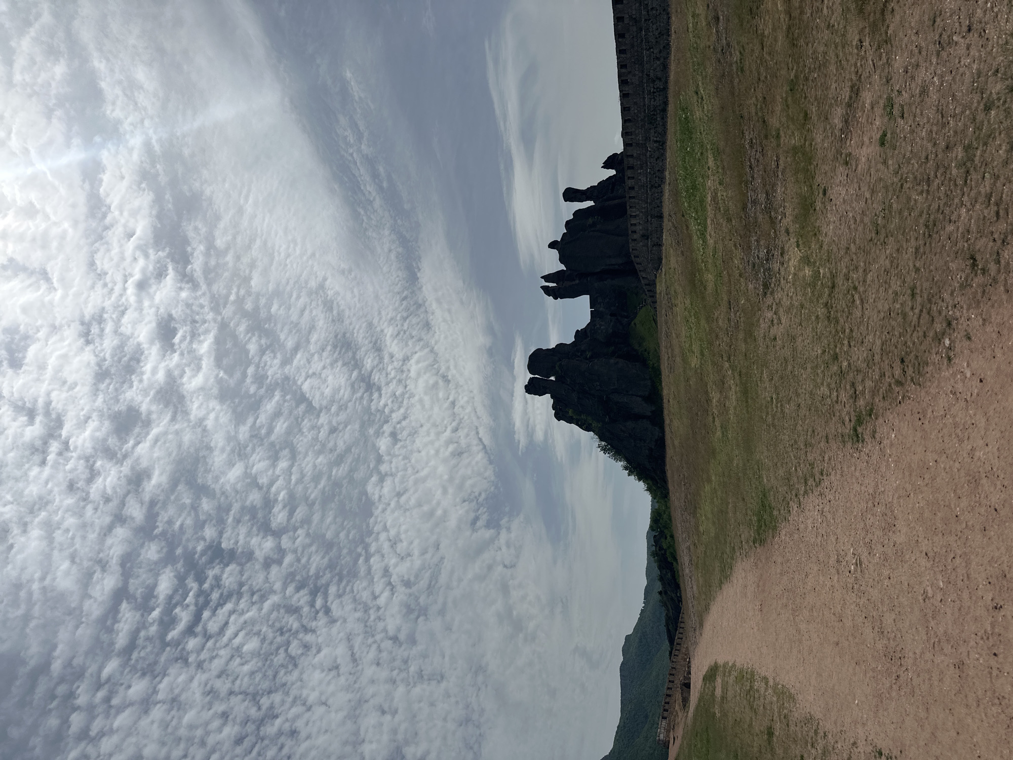 The image presents a landscape in Belogradchik, Bulgaria, featuring a prominent rock formation, part of the Belogradchik Rocks, under a sky filled with altocumulus clouds. A stone wall runs along the edge of the rock formation, while the ground is covered in dry grass and a dirt path. In the distance, a mountain range is visible.