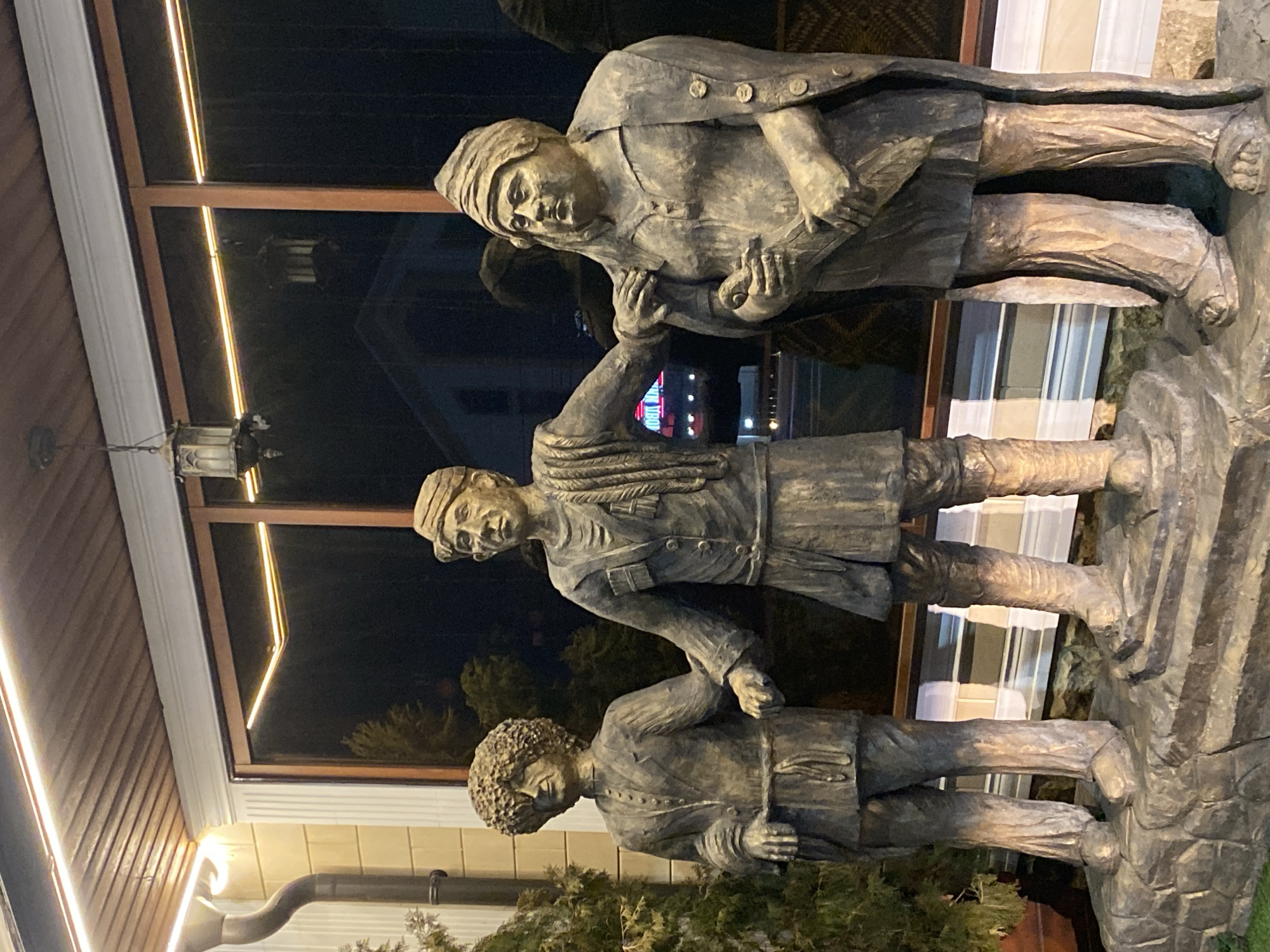 The image shows a sculpture of three men in a city of unknown location. The sculpture is positioned in front of a building with a window reflecting the night sky. The men are dressed in similar clothing, potentially indicating a historical or cultural connection. The sculpture's material appears to be stone or a similar durable substance. The surrounding environment includes vegetation and architectural details, suggesting an outdoor setting.