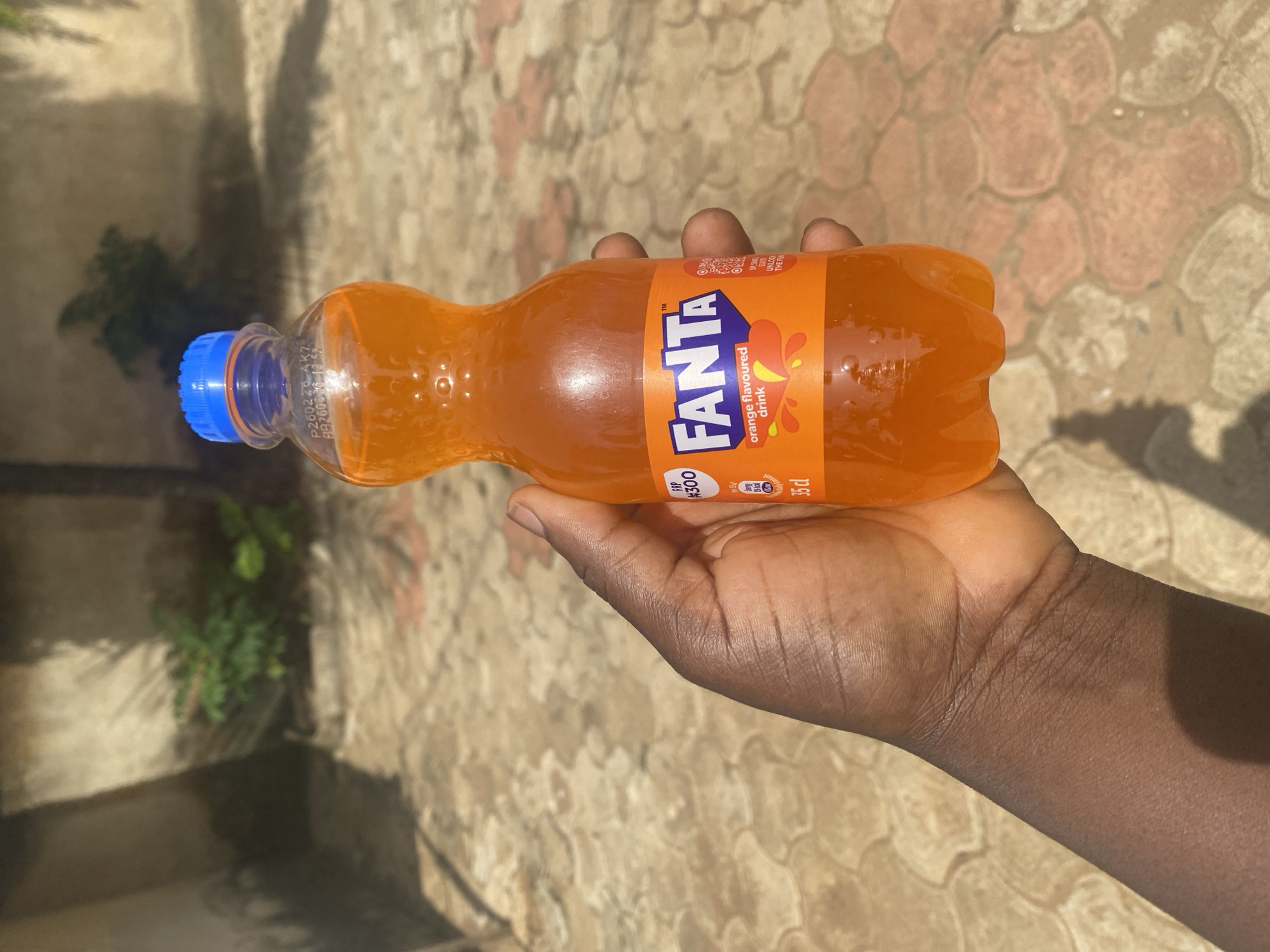 A person with dark skin is holding a 35 cl bottle of Fanta orange flavoured drink in their hand. The bottle has a blue cap. The background is a paved area with plants visible in the shade. The image was taken in an outdoor location in Maigizo, Nigeria.
