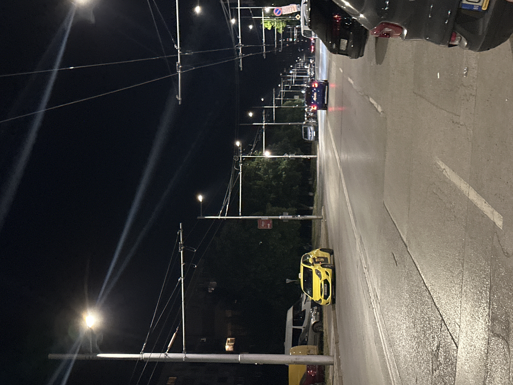 The image captures a night scene on a city street with trolleybus wires overhead. In the foreground on the right, the back of a dark-colored car is visible, parked on the side of the road. The car has a license plate that appears to be white with blue and yellow markings. Further down the street, a blue car is driving away from the camera, its taillights illuminated. On the left side of the street, a bright yellow taxi is parked at the curb. Behind the taxi, a white van and another car are also parked. The street is illuminated by streetlights, casting beams of light across the asphalt. Trees line the side of the road, and buildings are visible in the background. The overall impression is of a quiet urban street at night.