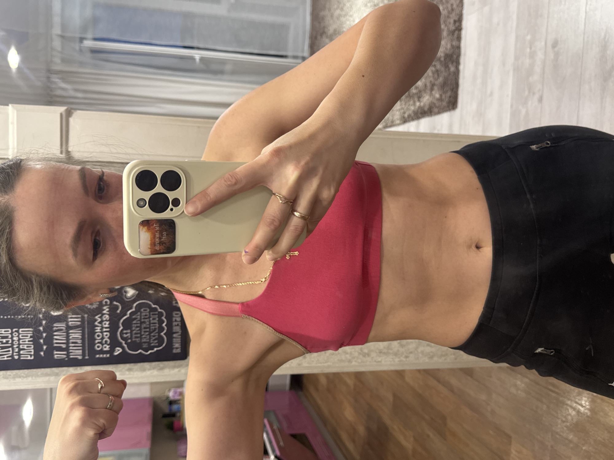 In an indoor setting in Chaikovsky, Russia, a woman is captured taking a mirror selfie. The image is rotated, showing her from the torso up. She is holding a light-colored smartphone with a prominent camera array in her right hand, positioned to reflect herself in the mirror. She is wearing a pink sports bra or crop top and black athletic pants or leggings. Her left arm is bent, possibly showcasing her arm definition, with a small gold ring on her pinky finger and another on her index finger. On her right hand, two more gold rings are visible: one on her ring finger and another on her pinky. A delicate gold chain with a cross pendant hangs around her neck.
The background reflected in the mirror reveals a room with white wall molding or trim, wooden flooring, and a textured rug. Prominently featured on the wall behind her is a dark surface, possibly a chalkboard, with extensive white, mirrored text. The OCR of this text includes fragments such as "درول", "OPEMVINY", "LIOTEN VAN", "ORPLINYN", "TWINLP", and "CWSHIPCH", among others, all appearing reversed due to the mirror reflection. The lighting suggests it is daytime or a brightly lit interior. The woman's expression is neutral, focused on the act of taking the photo, possibly documenting her fitness progress given her attire and pose.