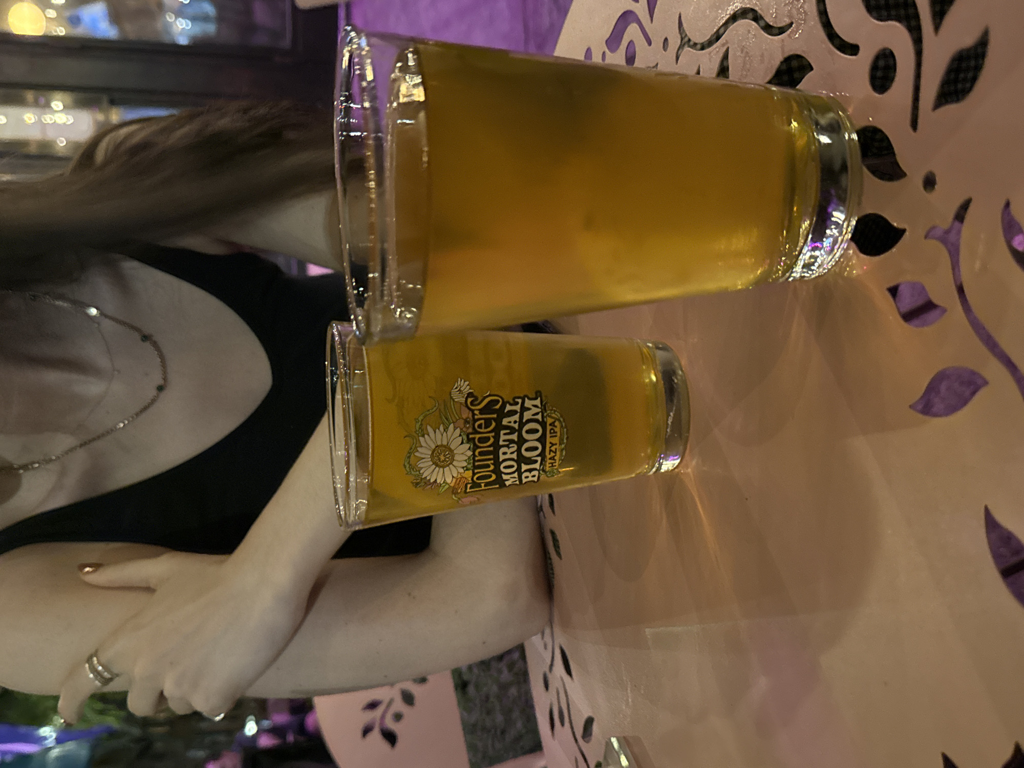 The image captures an indoor scene, possibly a bar or restaurant, located in New York City, United States of America. The focus is on two glasses of beer, presumably craft beers, positioned on a light pink table with decorative laser-cut patterns. The smaller glass displays a label indicating "Founders Mortal Bloom Hazy IPA". A woman is present in the scene, with her arm and hand visible. She wears a black top and a silver necklace. A ring is visible on her finger. The ambient lighting is dim, suggesting an evening setting. The background shows blurry lights, possibly from other establishments.