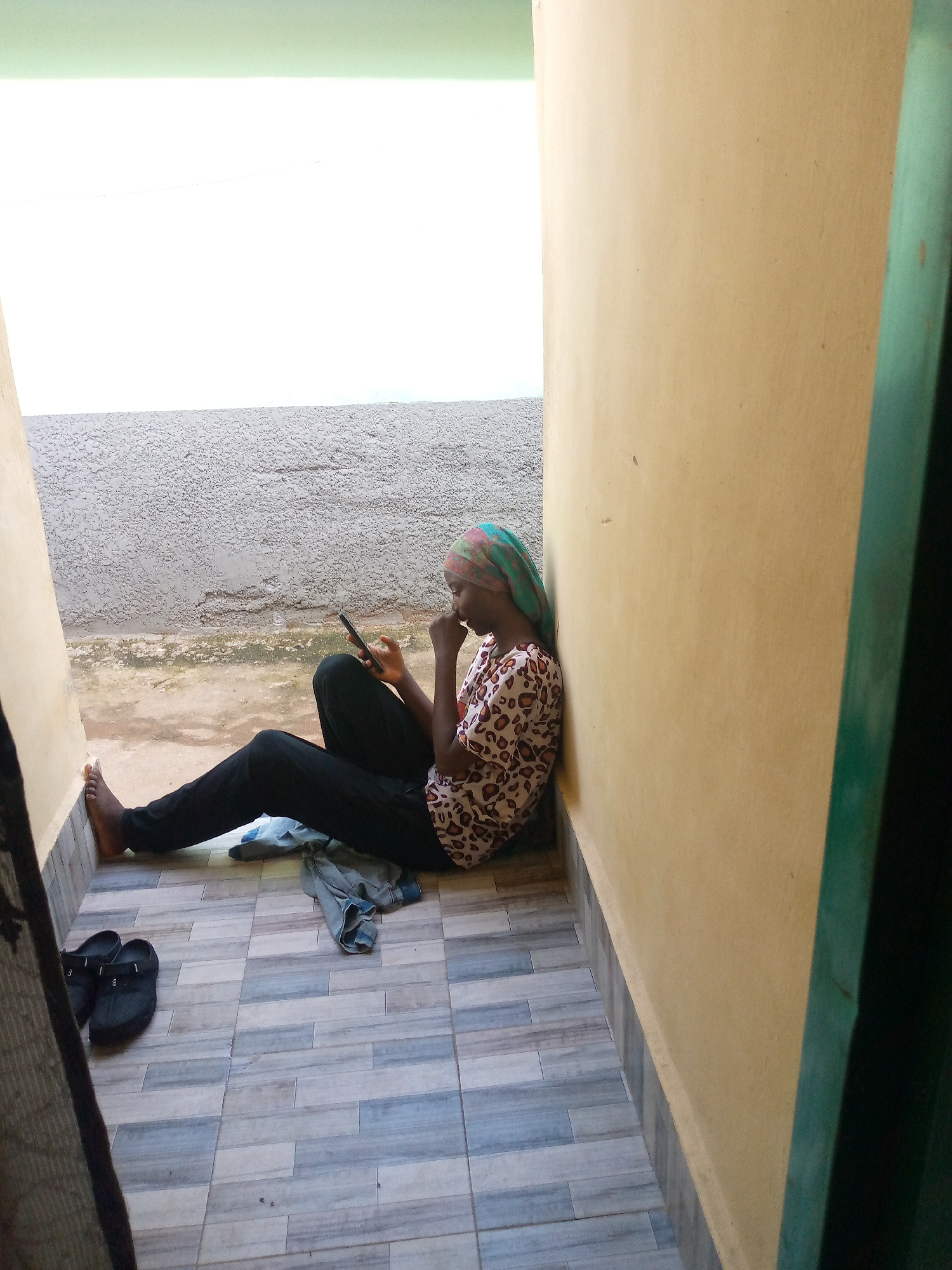 A young woman with a green and pink headscarf sits on a tiled floor, leaning against a light yellow wall. She is wearing a white shirt with a brown leopard print pattern and black pants. She is holding a dark-colored smartphone in her right hand and appears to be looking at it. Her left hand is near her mouth, with her thumb touching her chin. To her left, on the tiled floor, are a pair of black slip-on shoes and a piece of folded gray fabric, possibly clothing. The floor is tiled with a pattern of light and dark gray rectangular tiles. Behind her, there is a textured gray wall. To the left of the gray wall, a bright white surface is visible, and above that, a pale green band runs horizontally. The scene is outdoors, possibly on a balcony or patio, in Jalingo, Nigeria. The light suggests it is daytime.
