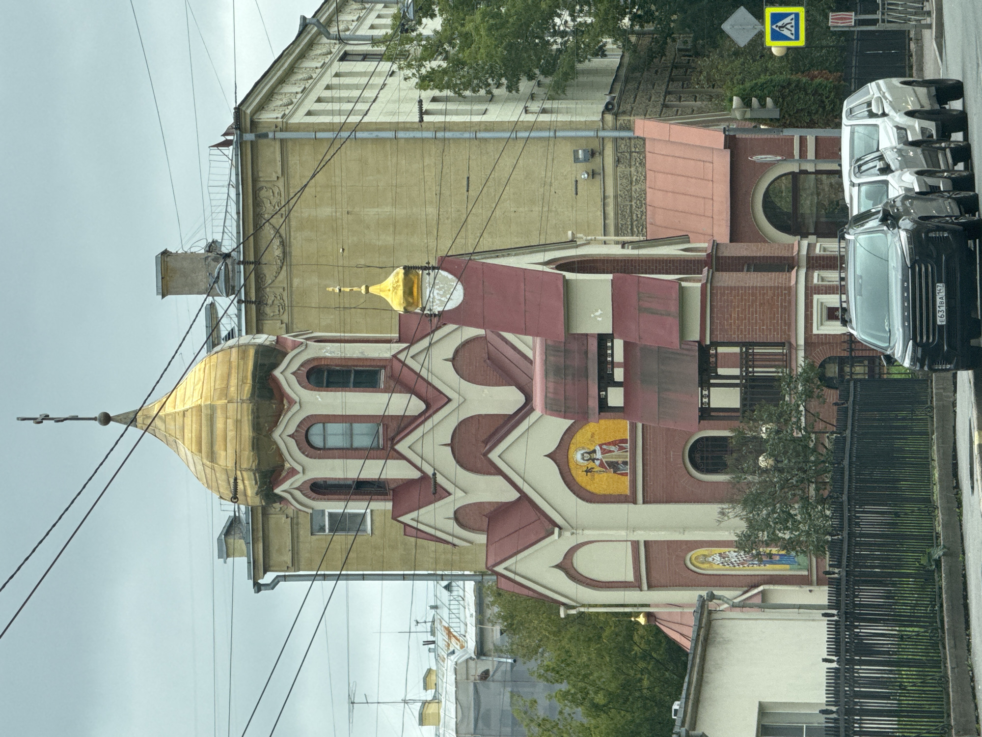 The image depicts a street scene in Saint Petersburg, Russia, featuring an Orthodox church with a prominent golden dome and red roof. The church's facade is decorated with mosaics. In the foreground, several cars are parked along the street, and a black metal fence runs along the sidewalk. Behind the church, larger buildings with yellow and white facades are visible. Overhead, electrical wires crisscross the sky, which is overcast. A pedestrian crossing sign and a traffic light are present on the right side of the frame. The overall impression is a typical urban setting with architectural elements of religious and residential buildings.