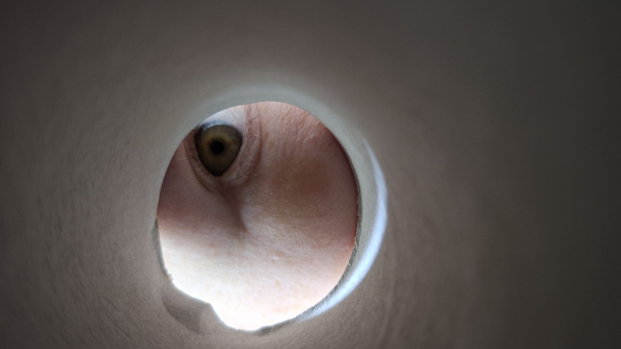 A close-up image shows a human eye peering through a circular opening. The eye is hazel in color, and the surrounding skin is light-toned. The opening appears to be in a light-colored, possibly paper or cardboard material. The environment is out of focus. There is no indication of the location being Большой Букор, Russia, or any other specific city.