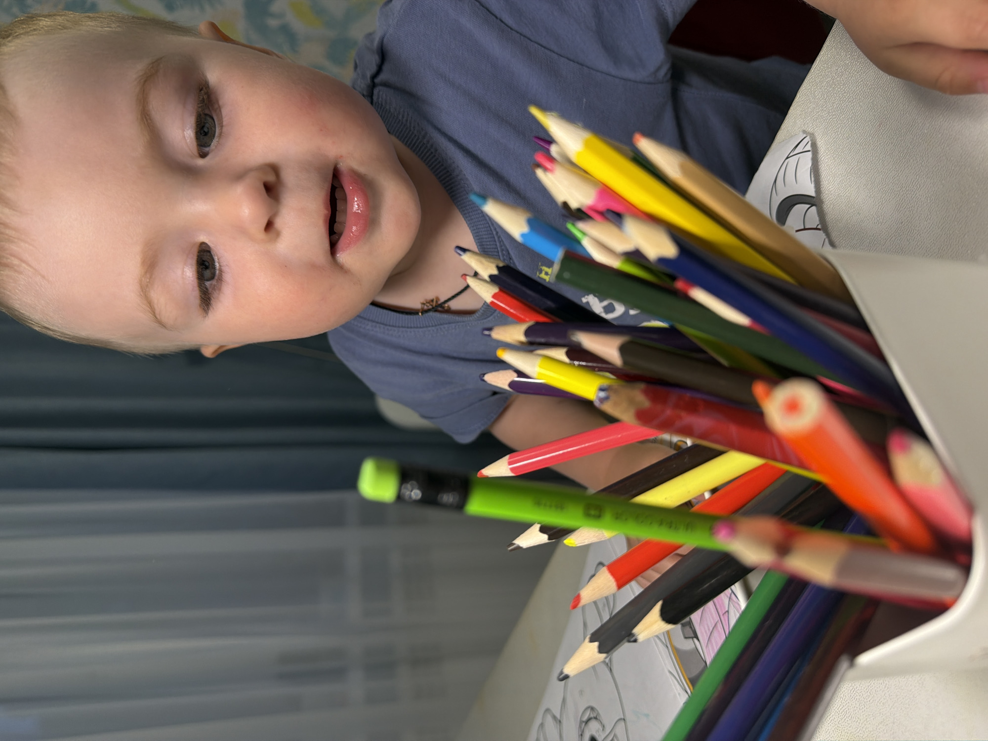 A young Caucasian child, appearing to be approximately 2-3 years old, with fair skin, light brown hair, and blue eyes, is positioned in the foreground of the image. The child is wearing a blue t-shirt and looking downwards and to the right, with their mouth slightly open, suggesting engagement or speech. Their expression is one of mild concentration or curiosity.
In front of the child, a white container filled with numerous colored pencils is prominently displayed. The pencils are of various colors including green, red, yellow, blue, black, and purple, with their tips pointing upwards. One green pencil in the center of the frame is in sharp focus, showing a black eraser tip.
The background reveals a textured grey surface that appears to be a table, on which a piece of paper with an illustration, possibly a drawing, is visible beneath the pencils. Further back, a blurred window with sheer curtains and vertical blinds is discernible. The overall lighting suggests an indoor setting, possibly a home environment. The image's composition focuses on the child and the art supplies, hinting at an activity of drawing or coloring.
The provided metadata indicates the location as Votkinsk, Russia. This context suggests the scene is situated within a residential or educational space in Votkinsk.