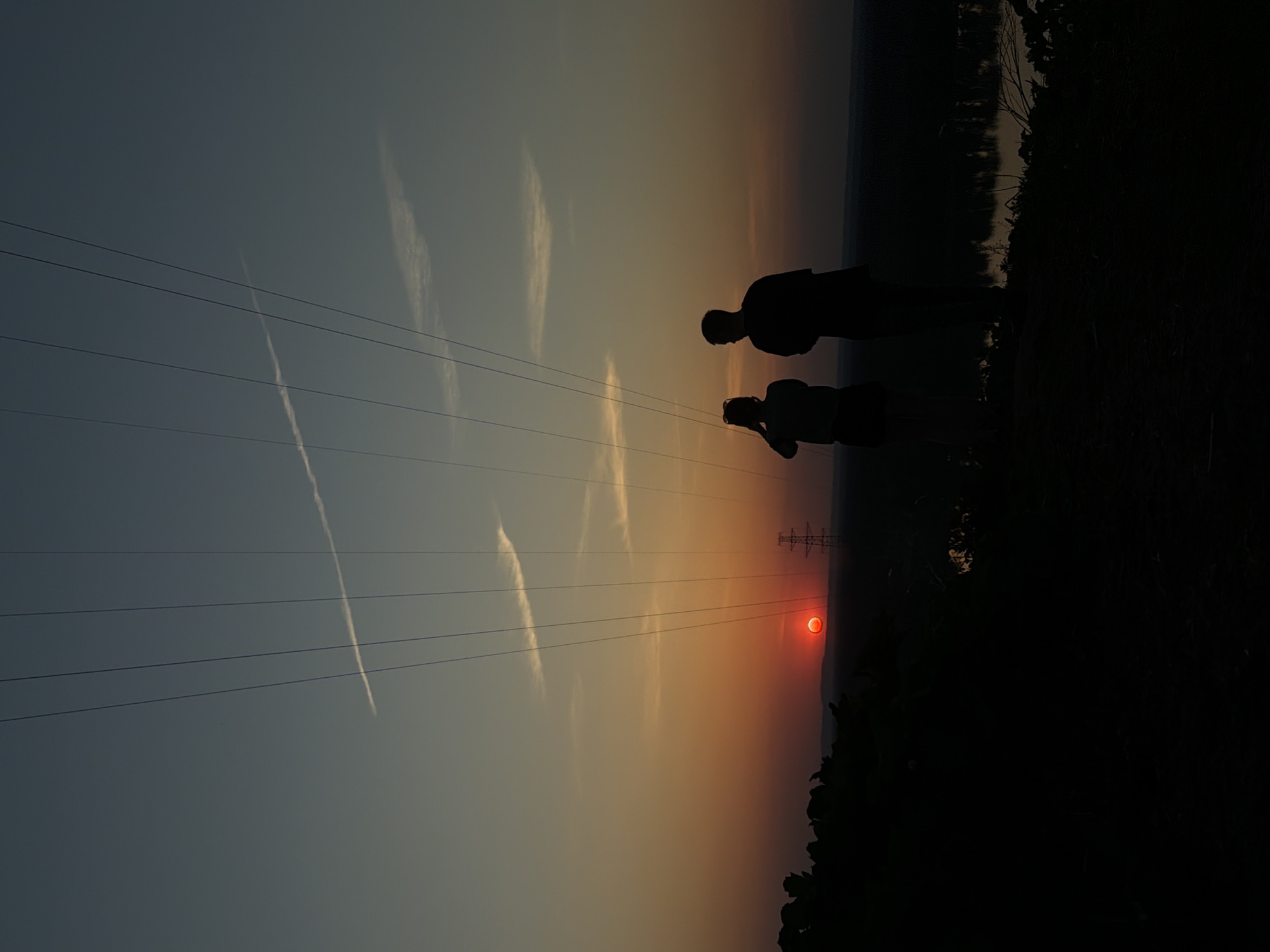 The image shows a sunset over the city of Noviy, Russia. Two silhouetted figures stand on a dark foreground, appearing to be a man and a woman. The woman is holding a camera, possibly photographing the sunset. A power line tower is visible near the setting sun. Power lines stretch across the image. The sky shows contrails and thin clouds. The landscape at the far right is dark.