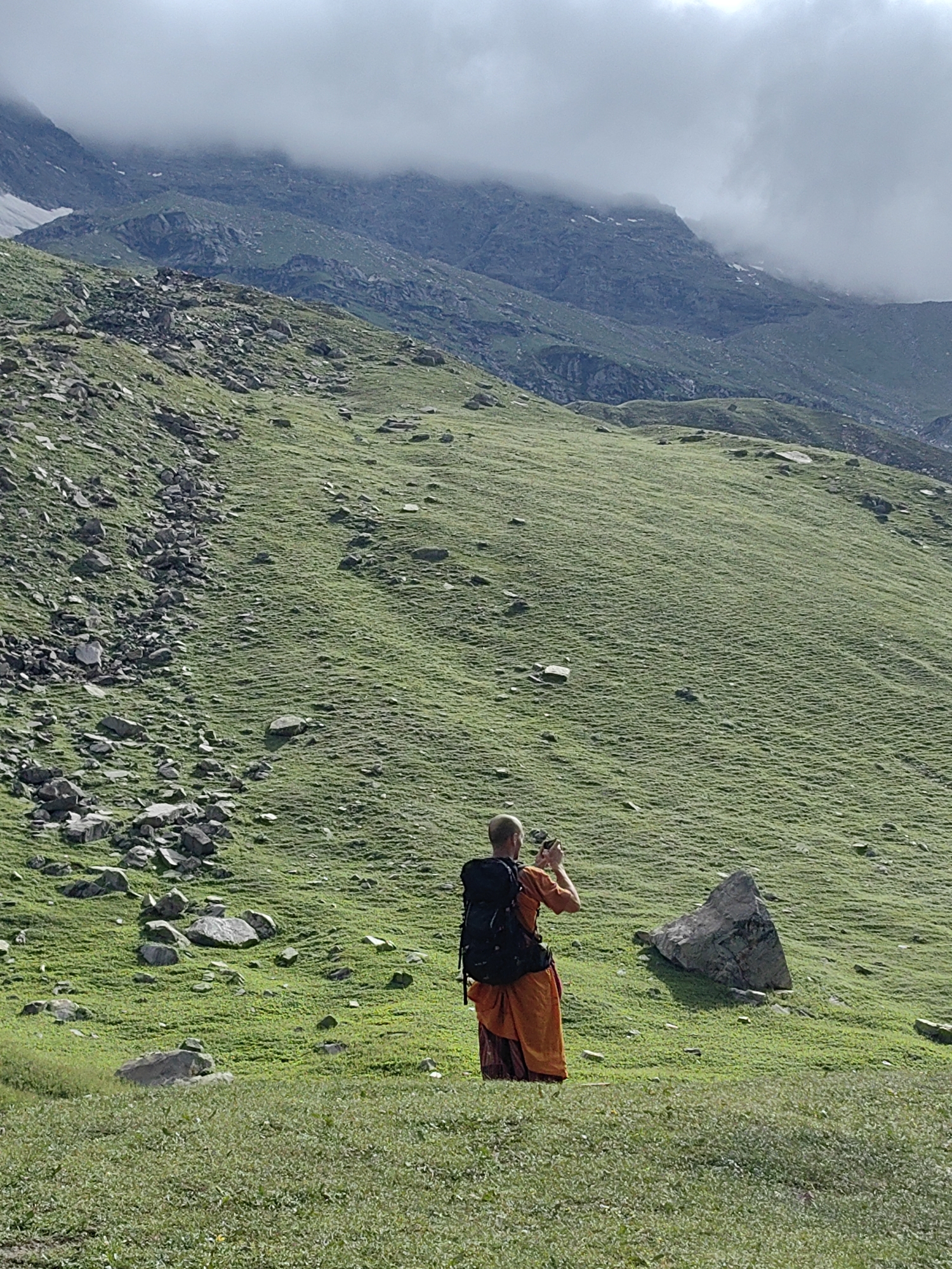 A person wearing an orange robe and a black backpack stands in a grassy mountain landscape. The person is facing away from the camera and appears to be taking a picture with a mobile phone. The background consists of a steep, green mountain slope dotted with rocks, with a cloudy sky and distant mountain peaks visible. The image is taken outdoors in what is likely a rural or mountainous area, possibly near the city of Bashisht, India, given the context. The ground in the foreground is covered in short green grass.