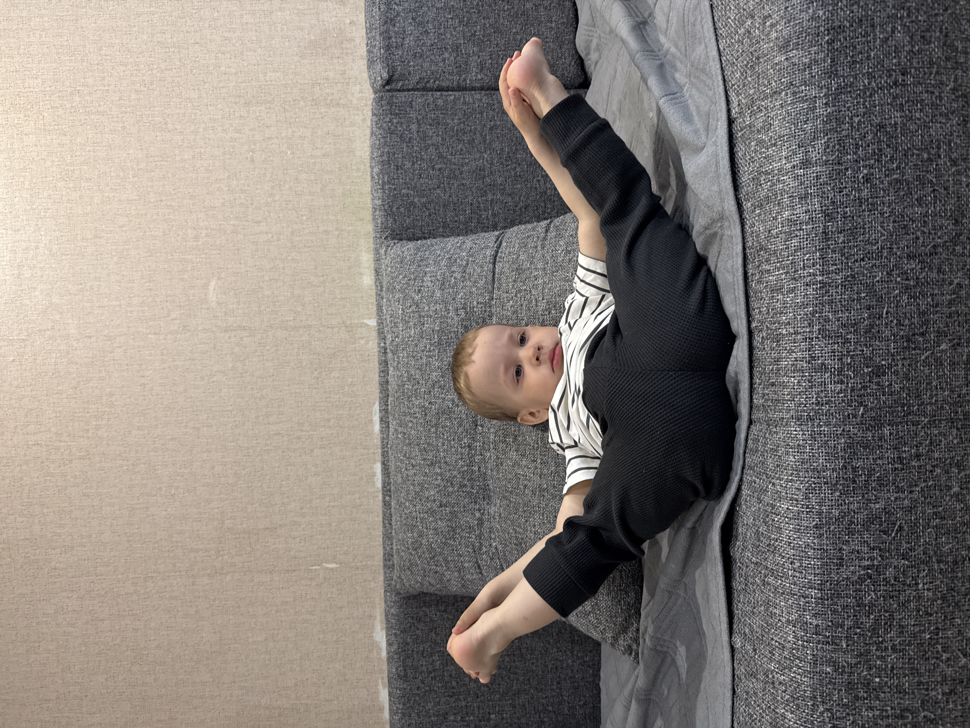 A young child is lying on a gray sofa, with their legs raised in the air. The child is wearing black pants and a striped shirt. The sofa is in front of a beige wall. The location is Rusanovo, Russia.