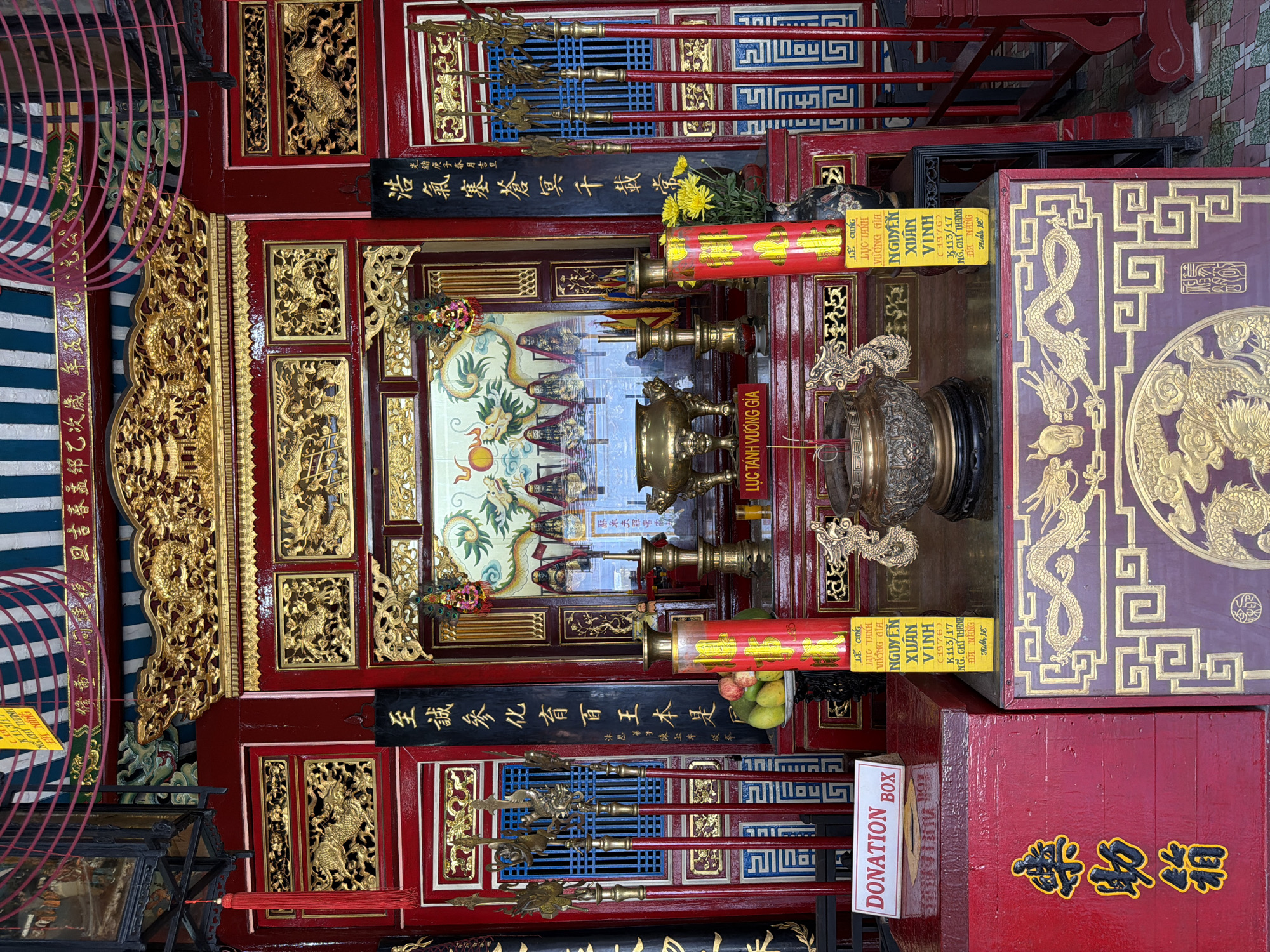 The image shows an ornate altar inside a building in Hội An Ward, Vietnam. The altar is elaborately decorated with red and gold, featuring intricate carvings of dragons and other traditional motifs. In the center of the altar, there is a large brass incense burner, surrounded by various religious offerings and decorative items, including yellow flowers and a plate of fruit. Behind the incense burner, a framed image depicts several figures in ceremonial attire. Flanking the central display are tall, red cylindrical objects with yellow labels, likely candles or offerings. To the left, a red box with the words "DONATION BOX" is visible. Vertical banners with Chinese or Vietnamese inscriptions hang on either side of the altar. The overall impression is one of a sacred and richly adorned space dedicated to religious worship.