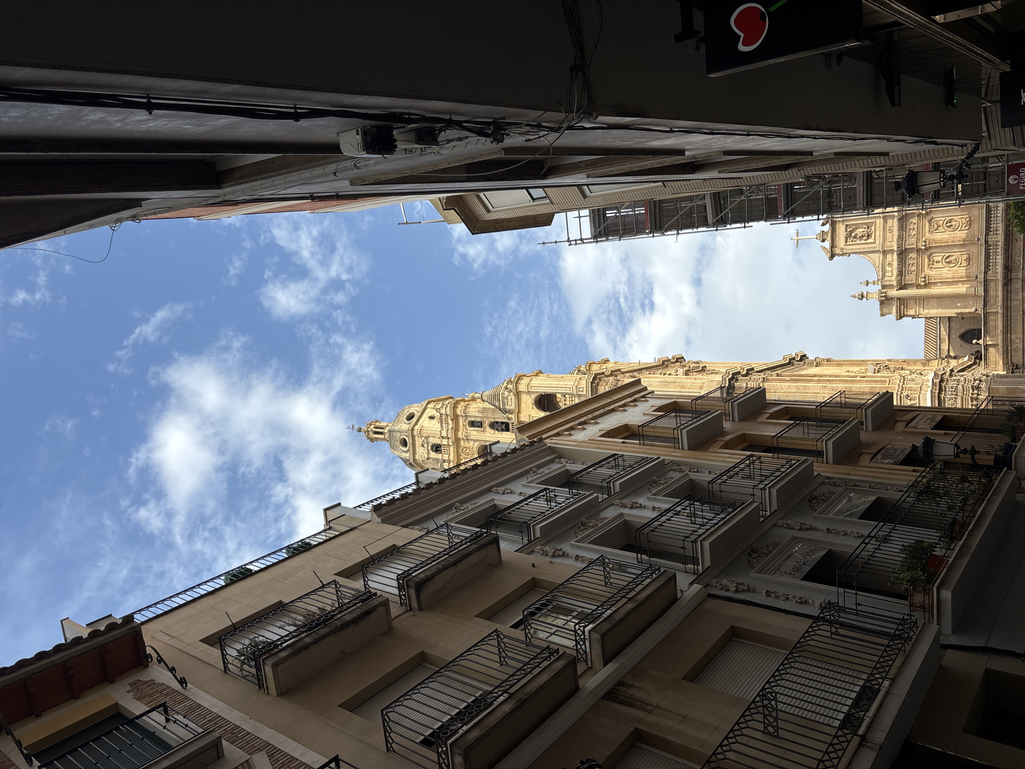 The image captures a narrow street in Murcia, Spain, looking upwards towards the sky. On the left, a building with multiple floors and balconies with wrought-iron railings lines the street. The building facade is a light tan color with some brick accents and window shutters. To the right, another building is partially visible, appearing darker and more modern, with visible electrical conduits and wiring on its exterior.
Dominating the background is the impressive Murcia Cathedral. Its intricate, golden bell tower with a dome is clearly visible, reaching towards the sky. Further into the background, the facade of the cathedral's main structure is visible, featuring a large cross atop its roof. Scaffolding is erected on a part of the cathedral, suggesting ongoing restoration or construction work.
The sky above is a bright blue, dotted with fluffy white clouds. The perspective is from street level, looking up, creating a sense of height and the grandeur of the architecture. There are no people visible in the frame. The overall scene depicts an urban environment characterized by historic architecture and a clear sky.