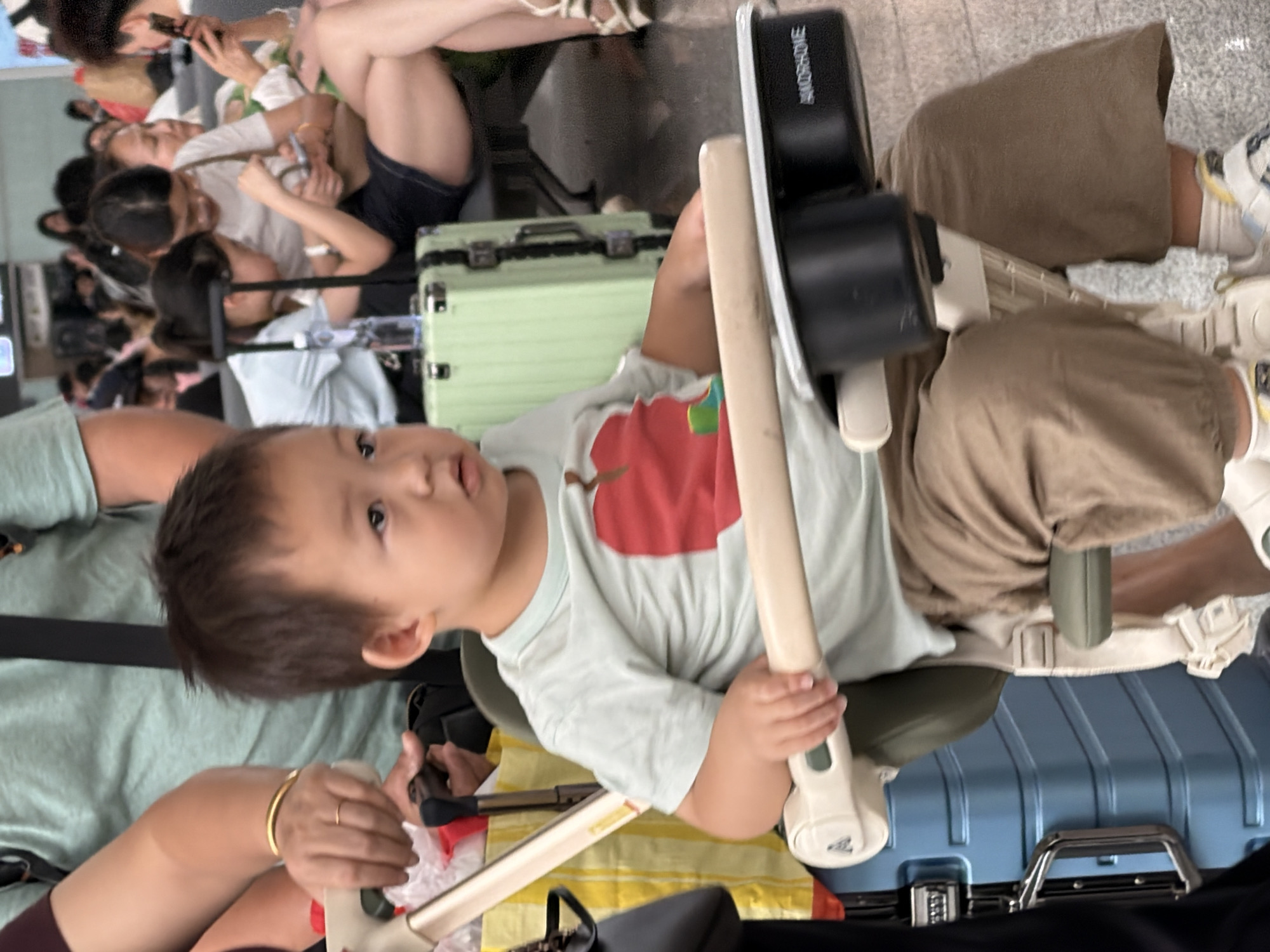 In Panyu District, China, a young child sits in a stroller at what appears to be an airport or train station waiting area. The child, facing slightly left, wears a light blue t-shirt with a red apple graphic and brown pants. They are holding onto the stroller's tray. The stroller itself is white and green with black cup holders attached. Behind the child, a blue suitcase with a silver handle is visible, along with a light green suitcase. In the background, other people are seated and standing, some interacting with their phones. The overall environment suggests a public transportation hub.
