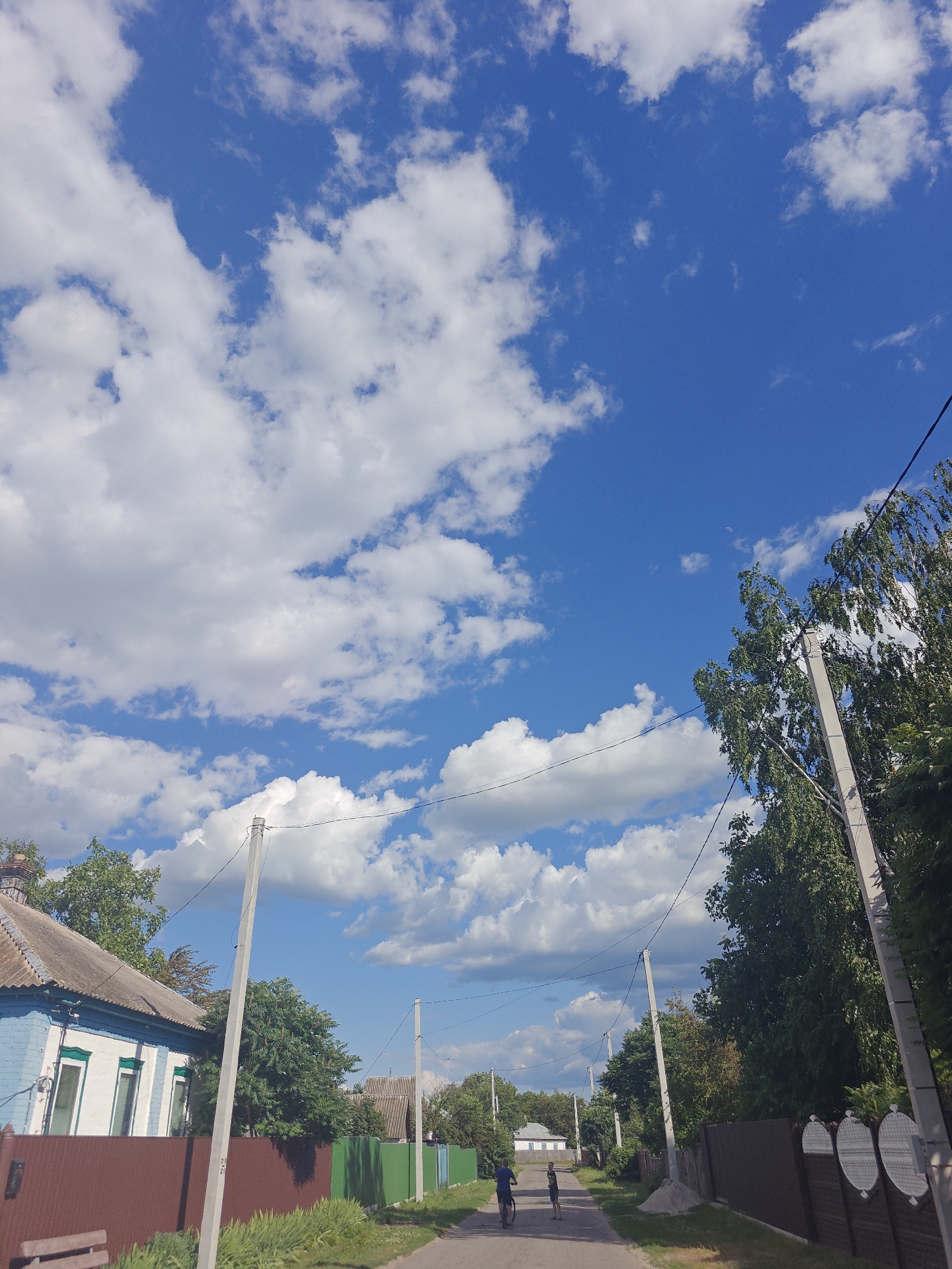 The image depicts a street scene in Kotelva, Ukraine, under a bright blue sky with scattered white clouds. On the left, a two-story house with a blue and white facade and a grey roof is visible. A brown fence runs along the front of the property. A rural road stretches into the distance, lined with trees and fences. Two individuals are present on the road: one is riding a bicycle, and the other is standing nearby. Several utility poles with wires are also present along the roadside. The overall impression is that of a quiet, residential area on a sunny day.