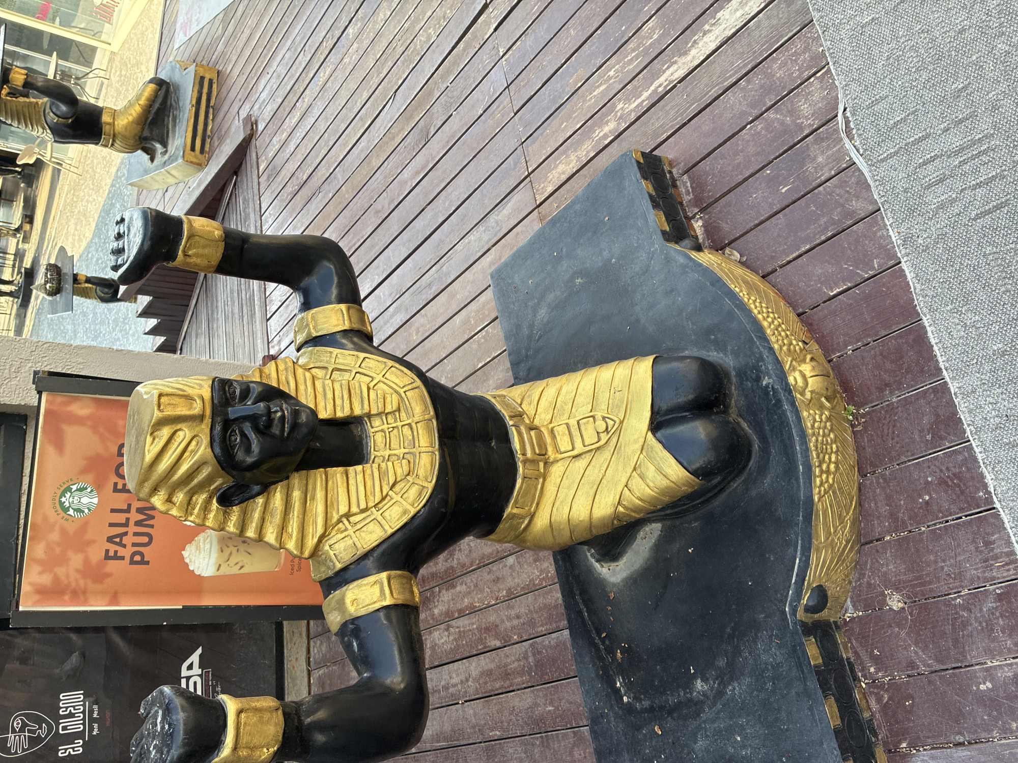 This image shows a statue of an Egyptian pharaoh kneeling with its arms raised in the city of Alanya, Turkey. The statue is black with gold accents on its headdress, collar, armbands, and kilt. The statue is positioned on a black platform with gold trim, which is placed on a wooden deck. In the background, a sign displays "FALL FOR PUMPKIN" and a Starbucks logo. The area appears to be an outdoor space, possibly a promenade or plaza, with other statues and seating visible in the distance. The lighting suggests it is daytime, and the environment appears to be a commercial or tourist area.