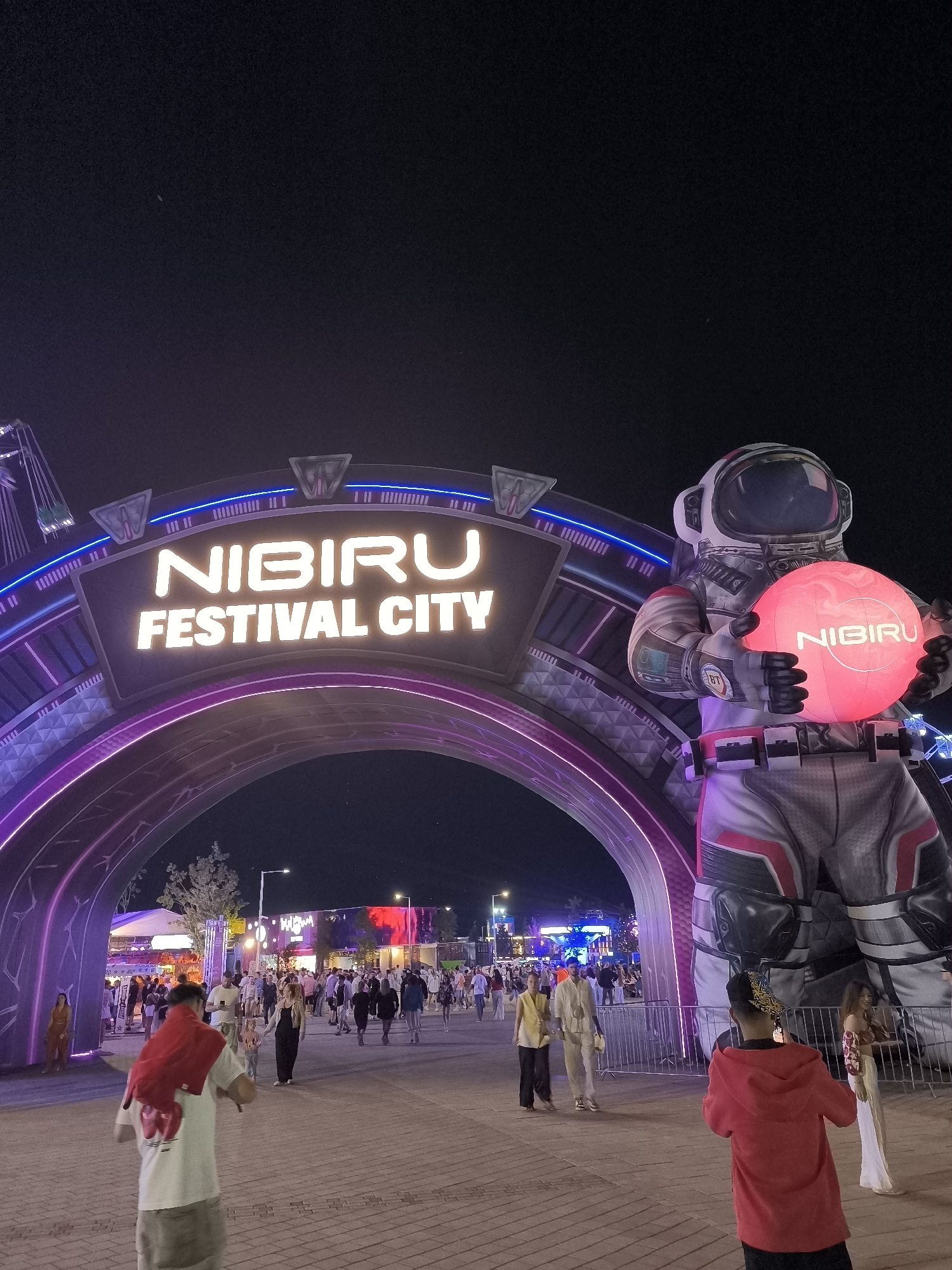 This is a nighttime, eye-level, vertical shot of the entrance to "Nibiru Festival City" in Costinești, Romania. A large, illuminated archway frames the entrance, with the words "NIBIRU FESTIVAL CITY" prominently displayed in glowing white letters. To the right of the archway stands a colossal inflatable astronaut figure holding a glowing pink sphere with the word "NIBIRU" printed on it. The astronaut is detailed with a white suit, silver accents, and red trim.
Beyond the archway, a crowd of people is visible walking through a paved area. Various stalls, structures, and lighting displays suggest a festival or entertainment venue. The ground is paved with cobblestones. The overall atmosphere is festive and illuminated by artificial lights against a dark night sky.