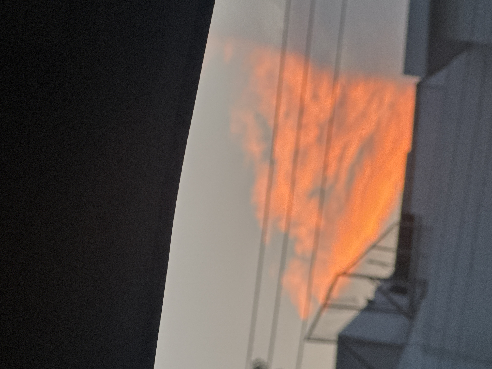 The image shows a partial view of a wall or building in the city of Puebla, Mexico, with a reflection of an orange sunset or sunrise. The reflection is likely cast by the windows on a nearby building and the scene is set against a light gray sky. Part of a black object, possibly a monitor, is seen in the left portion of the image. The wall exhibits vertical lines or markings. A portion of another building is visible on the right, showing metal beams or structures.