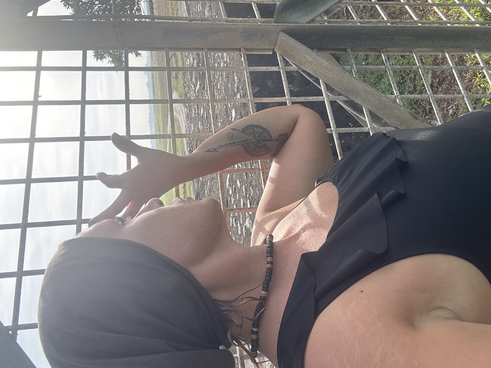 A woman lies on a metal grid structure outdoors. She wears a black garment and a beaded necklace. Her head is covered with a black scarf. She has a tattoo on her left forearm, which is raised towards her face. The background shows a landscape under a cloudy sky, visible through the grid. The location appears to be an open area with grass and possibly a road. It could be in Denpasar, Indonesia.
