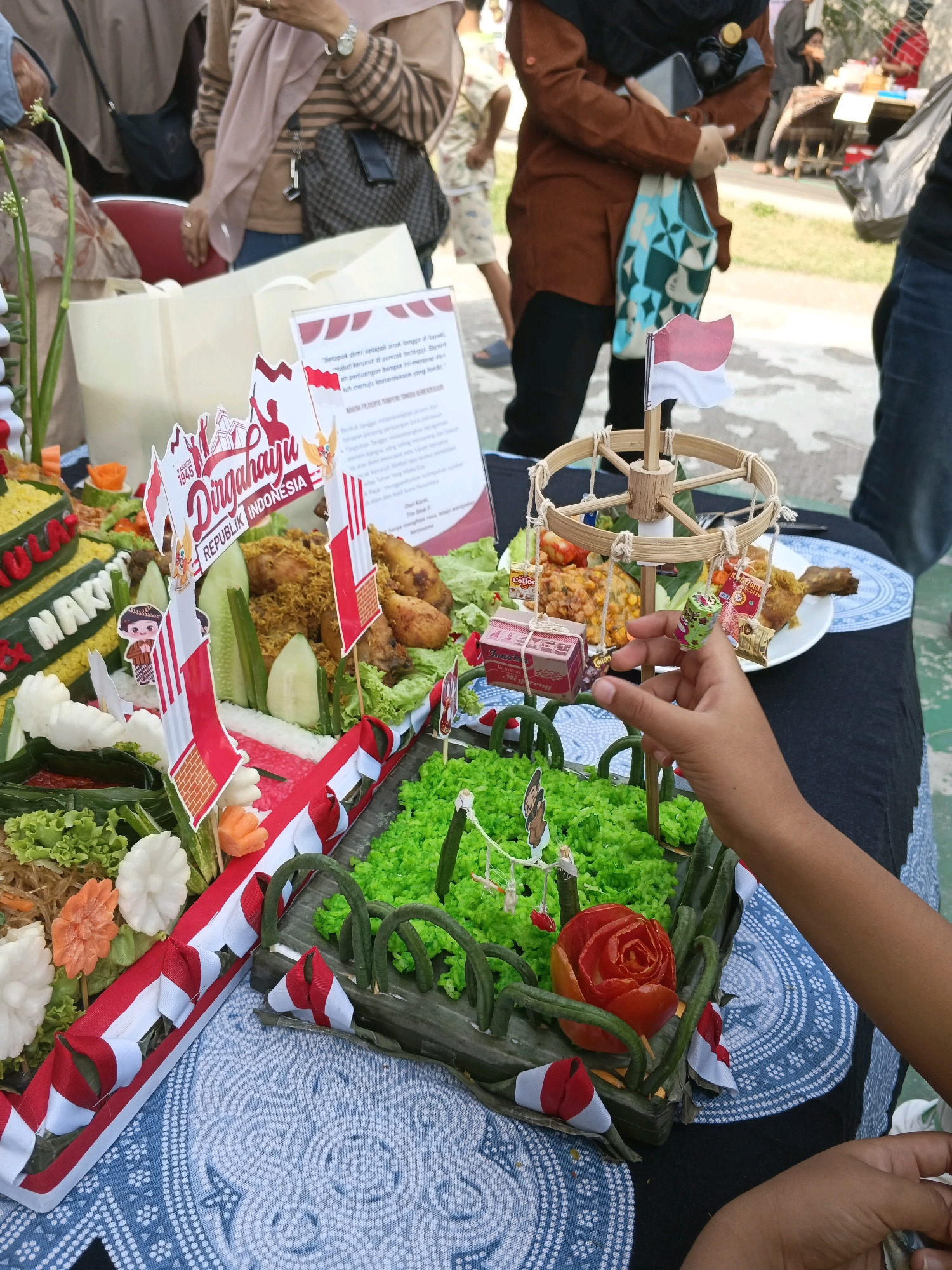 The image depicts a close-up view of a food display, likely part of an event or competition, at an outdoor market in Margahayu, Indonesia. In the foreground, a hand is reaching towards a small carousel-like decoration with hanging miniature items. The main subject is a colorful, tiered food arrangement, possibly a traditional Indonesian dish like Nasi Tumpeng, decorated with red and white Indonesian flags and miniature figures. The food appears to include rice, chicken, and vegetables. The background shows people milling around, with some holding bags and wearing casual clothing, suggesting a public gathering. The overall setting is outdoors, with a glimpse of grass and pavement.