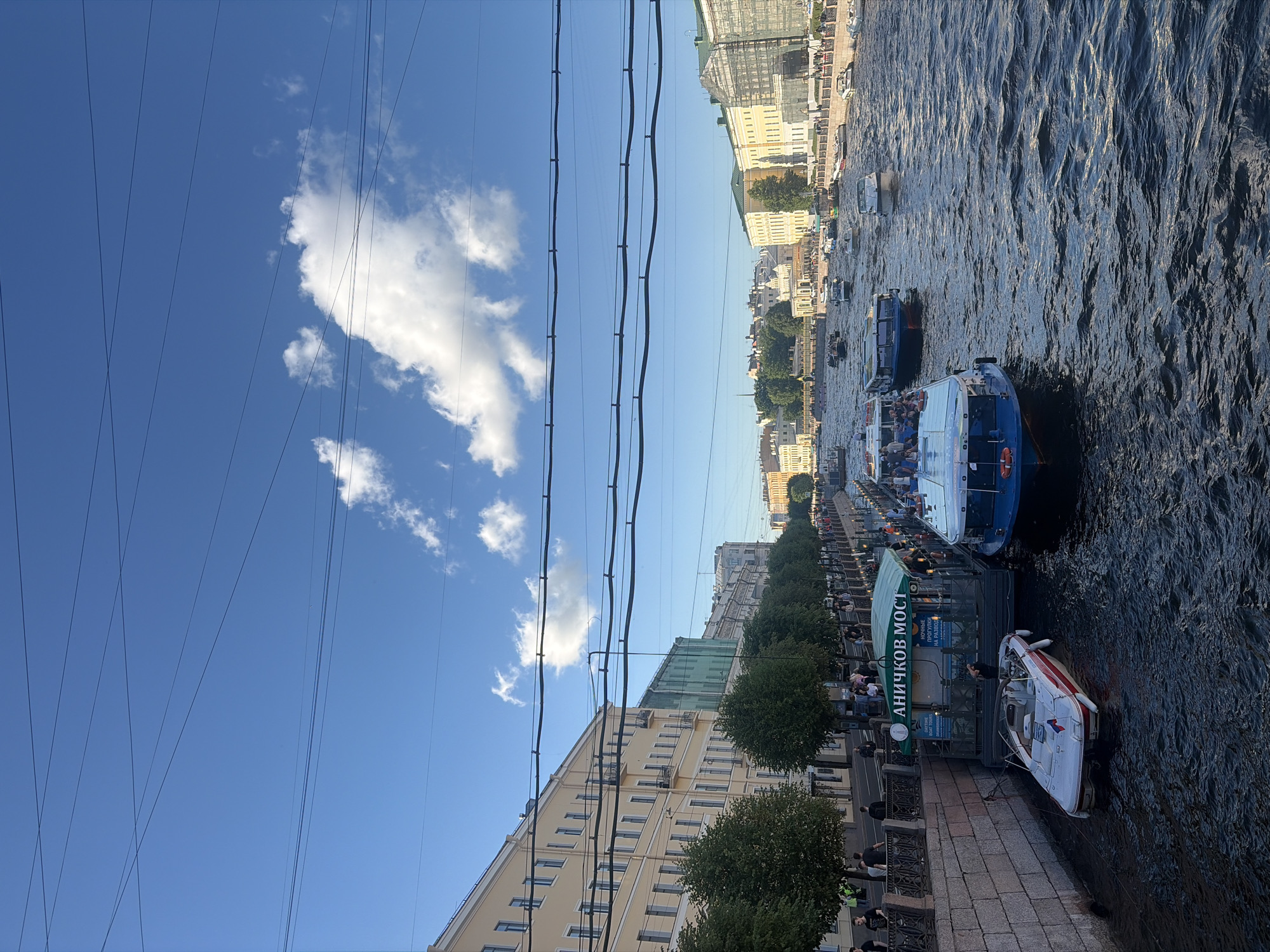 This image captures a scenic view of the Fontanka River in Saint Petersburg, Russia, on a bright, partly cloudy day. Along the riverbank, a row of light yellow, multi-story buildings with numerous windows lines the street, interspersed with green trees. The river itself is bustling with activity, featuring several tour boats and smaller vessels. In the foreground, a white and red speedboat is moored near a pier with a green awning that has "АНИЧКОВ МОСТ" (Anichkov Bridge) written on it in Russian, indicating boat tours are available. People are visible walking along the embankment and on the boats. The sky is a vibrant blue with scattered white clouds, and power lines crisscross the upper portion of the frame. The overall scene depicts a lively urban waterway scene with iconic St. Petersburg architecture.