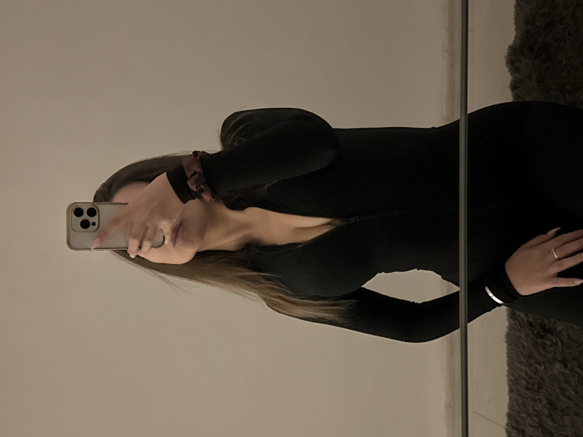 The image, originally oriented vertically but displayed sideways, captures a mirror selfie of a person, likely female, taken indoors. The subject is visible from the upper torso to approximately mid-thighs, standing in front of a mirror. They are wearing a form-fitting black long-sleeved top or dress, possibly a jumpsuit, with a front zipper that is partially open, revealing a hint of cleavage.
The person has long, straight, dark brown hair that falls over their left shoulder. Accessories include a dark red or maroon scrunchie on their left wrist, a silver ring on the ring finger of their right hand, and a thin silver bracelet on their left wrist. Their face is largely obscured by a light-colored smartphone, likely an iPhone, which they are holding with both hands to take the selfie. Only the chin, jawline, and parts of the lips are visible, suggesting a neutral or slightly posed expression.
The background reflected in the mirror behind the subject is a plain, light-colored wall, appearing off-white or light grey. To the right of the mirror's vertical frame, a dark, textured object, possibly a shaggy rug or a piece of dark fabric, is partially visible. The overall lighting is subdued, indicating an indoor setting with artificial illumination, possibly in the evening. There is no visible text within the image. While no specific architectural or geographical cues are present, the photo was taken in Sofia, Bulgaria.