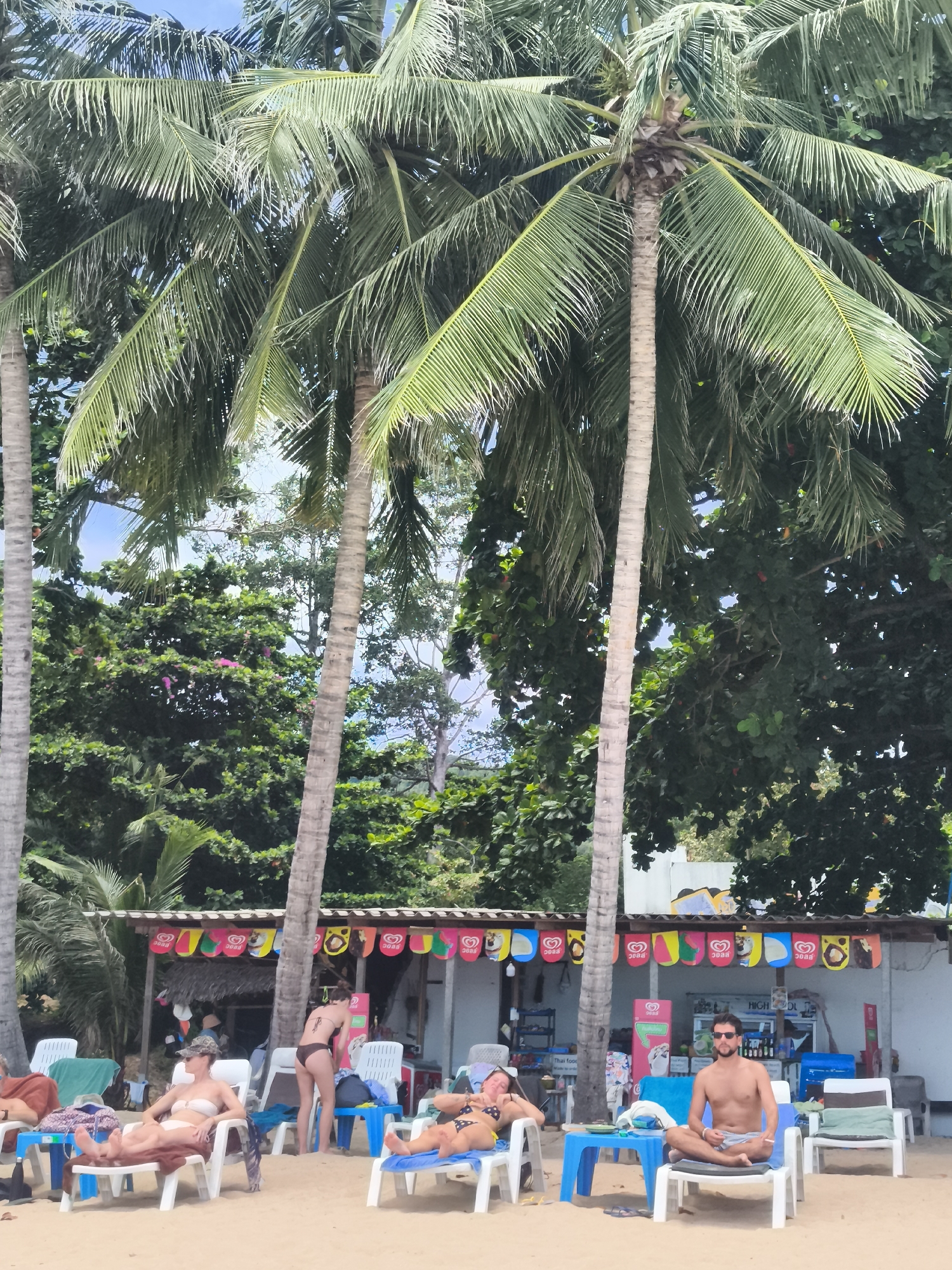 In the city of Ko Samui District, Thailand, an outdoor scene unfolds on a sandy beach. Palm trees with lush green fronds dominate the upper portion of the image, providing shade. Beneath the trees, a row of open-air stalls with colorful awnings serves as a backdrop. Several people are visible relaxing on the beach.
In the foreground, a man is seated cross-legged on a white lounge chair in front of a blue table, wearing sunglasses and light-colored shorts. To his left, a woman reclines on a similar lounge chair, wearing a dark bikini. Further to the left, another woman is seated on a lounge chair, wearing a white bikini top and bottoms, with a patterned sarong draped over her lap.
In the middle ground, a woman wearing a brown bikini stands near the stalls, appearing to be looking down. Behind her, other individuals can be seen partially obscured. The stalls themselves display various colorful flags or banners, and some appear to be selling refreshments. White plastic chairs and blue tables are scattered across the sand. The overall environment suggests a relaxed beach setting.