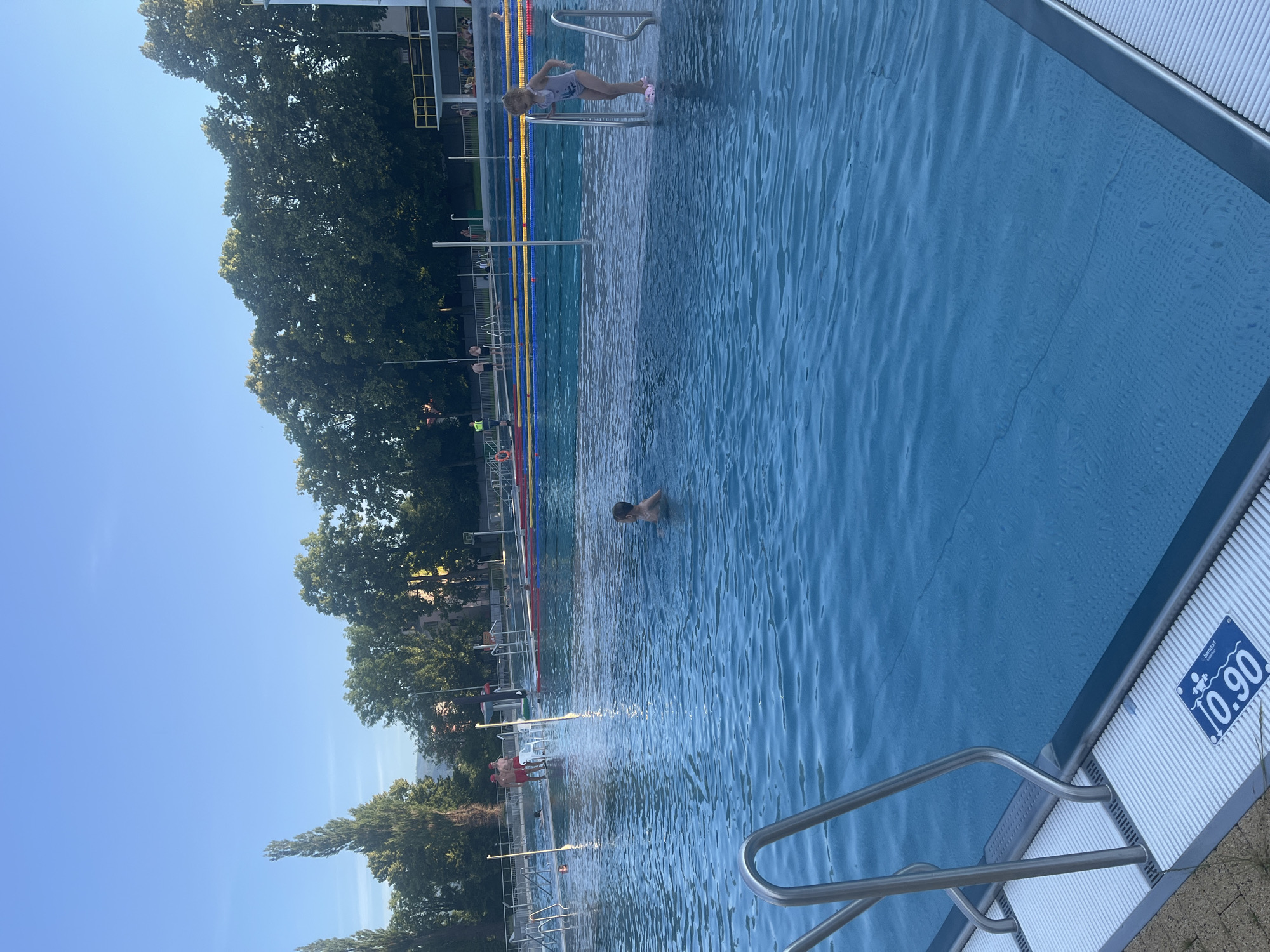 In the foreground of the image, the edge of a bright blue swimming pool is visible, with metal handrails and a white, textured surface leading into the water. A blue sign with white text indicating "10.90" is affixed to this surface. The water is clear and rippled, with the pool floor visible in some areas.
In the middle ground, a young boy with short, dark hair is swimming in the pool, facing towards the right of the frame. Further back, a young girl wearing a light-colored swimsuit is standing at the edge of a shallower section of the pool, holding onto a metal railing. Two adults, one wearing a red cap and red shorts, and another wearing a similar cap and red trunks, are standing near the edge of the pool.
In the background, the swimming pool extends further, with lane markers visible in the water. A diving platform with yellow railings is situated in the distance. Trees with lush green foliage line the perimeter of the pool area, and a few buildings are visible behind the trees. The sky above is a clear, bright blue, suggesting a sunny day. The scene appears to be an outdoor public swimming pool.