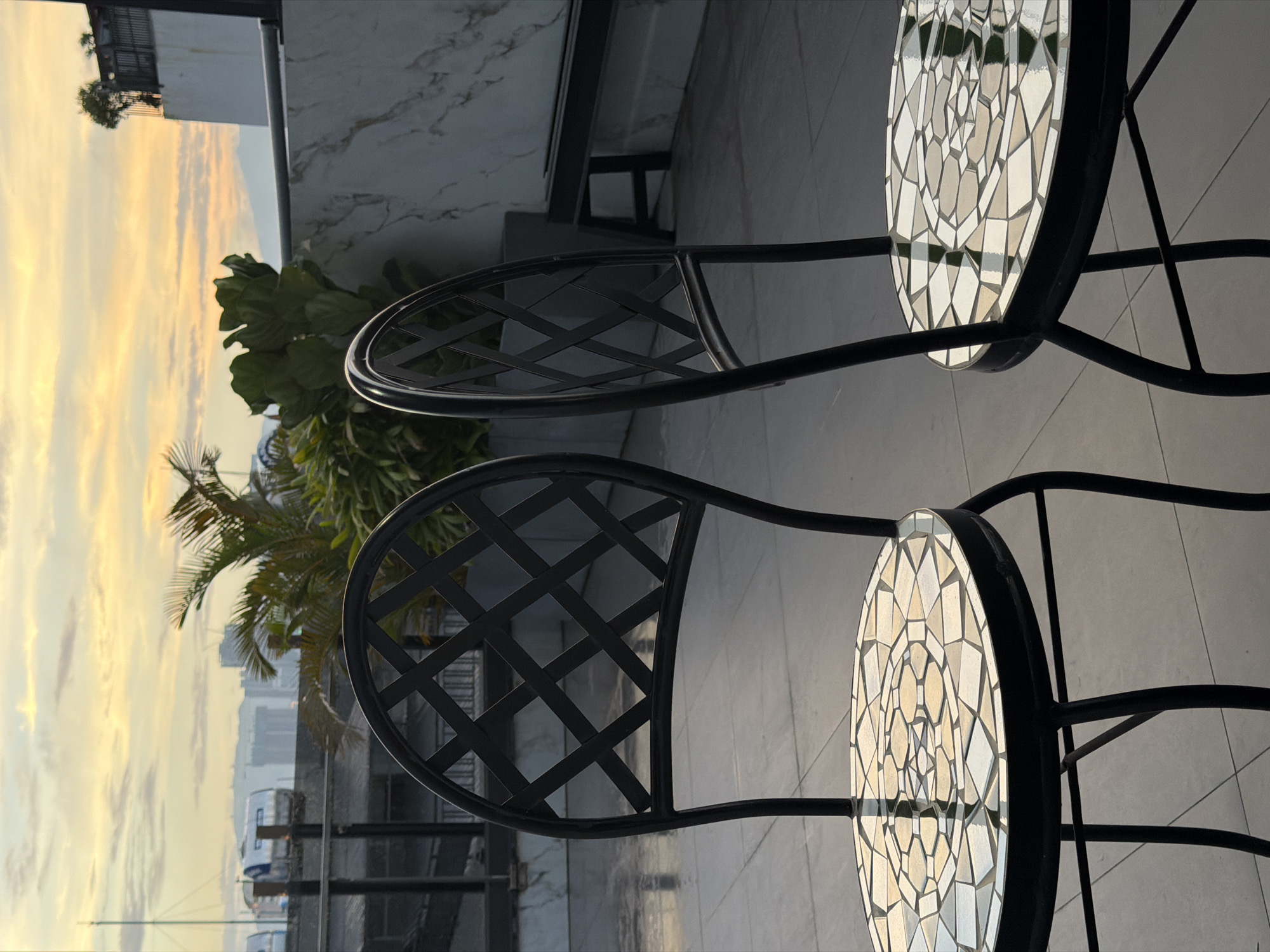 The image depicts an outdoor seating area, likely a rooftop or balcony, in Da Nang, Vietnam, during a sunset. Two black metal chairs with mosaic tabletops are prominently featured in the foreground, positioned on a tiled floor. The chair on the left is angled slightly towards the viewer, while the chair on the right is closer to the edge of the frame, showing its side profile.
Behind the chairs, a lush green plant with large leaves and palm fronds adds a touch of nature to the urban setting. The background reveals a hazy cityscape with tall buildings and mountains in the distance, characteristic of Da Nang. The sky is painted with the warm hues of sunset, with streaks of orange and yellow clouds. In the upper right corner, a white building with a marble-like facade and a black bench are visible, suggesting an area for relaxation or dining. The overall atmosphere is tranquil, with the soft light of the setting sun casting reflections on the wet tiled floor and the mosaic tabletops.