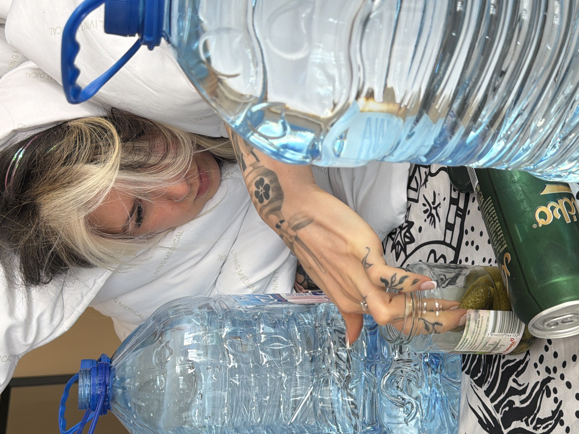 The image depicts a person lying on a white pillow or bedding, with part of their head and face visible. They have light blonde and black hair. A tattooed hand reaches towards a glass jar filled with pickles, which rests next to a green can of "Zlatko" drink on a patterned black and white surface. Two large bottles of water are positioned above and below the person, creating a visual frame. The backdrop includes the label "LIVING MEDICAL" on the white fabric. The image does not contain information to determine the location in the city of Budva, Montenegro.