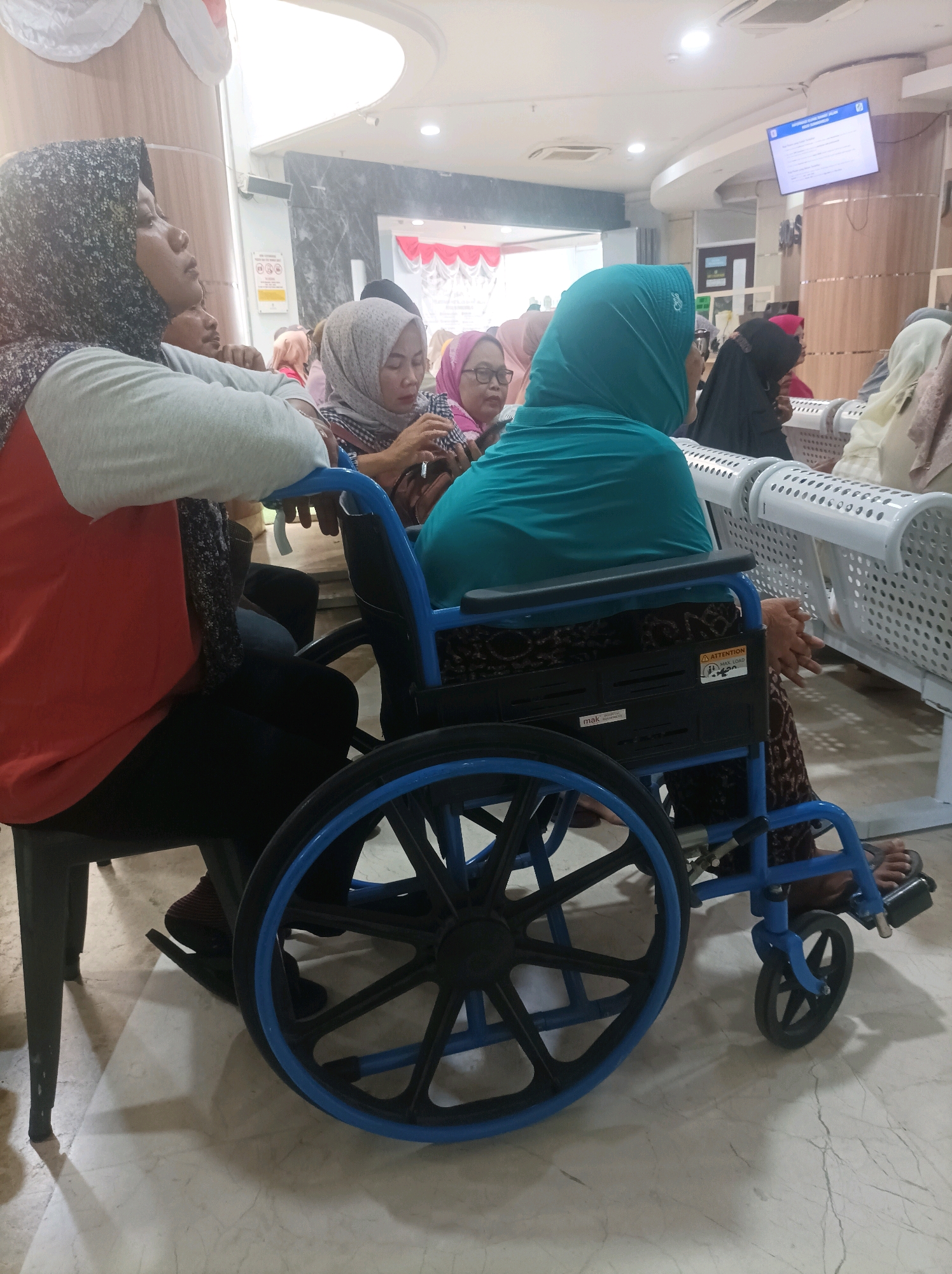 The image depicts a waiting area within a building, likely a clinic or hospital, in Sumuragung, Indonesia. In the foreground, a woman is seated in a blue wheelchair. She is wearing a teal colored headscarf and a patterned skirt. To her left, another woman is seated in a chair, wearing a grey headscarf and an orange shirt with a black patterned scarf draped over her shoulders. Her arms are crossed, and she appears to be looking upwards and to the left. Behind them, several other individuals are visible, some seated on benches and others standing. The environment features polished marble flooring, white metal benches with perforated seats, and a digital display screen mounted on the wall. The lighting suggests an indoor setting, possibly with overhead fluorescent lights. The overall scene conveys a sense of waiting or congregational gathering.