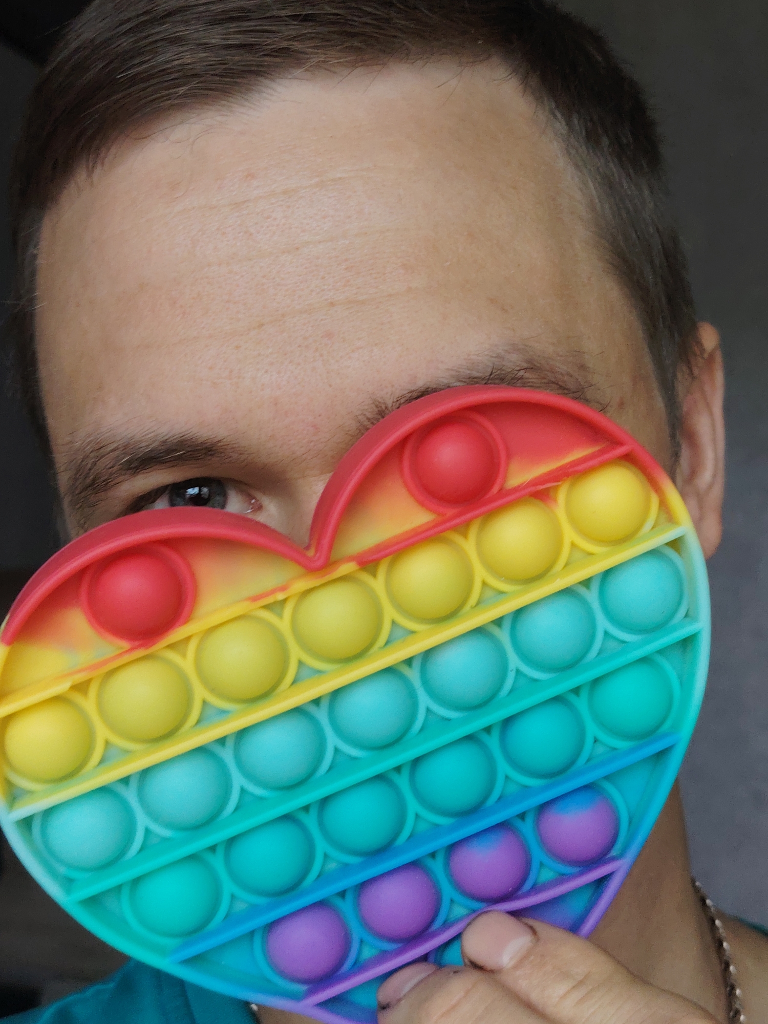 A person is holding a rainbow-colored heart-shaped "pop it" toy in front of their face, partially obscuring it. The person has light skin, short brown hair, and an eye visible behind the toy is light blue. The "pop it" toy is a gradient of colors from red at the top to purple at the bottom. The person's fingers are partially visible at the bottom corner of the toy. The person wears a green shirt and there is a thin silver necklace at their neck. The background is out of focus and dark gray. There is no information available to identify the location as the city of Chaikovsky, Russia, or any other specific location.