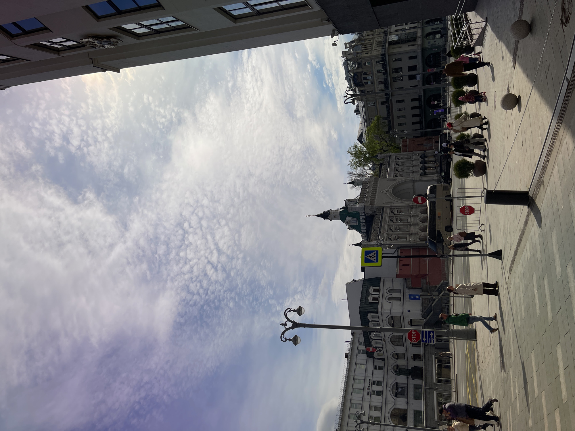 The image shows a street view in Moscow, Russia, with a clear sky filled with cirrocumulus clouds. Buildings line both sides of the street, with architecture ranging from traditional to modern styles. People are walking along the sidewalk, and street furniture such as lampposts and bollards are visible. A pedestrian crossing sign is present, as is a stop sign. The perspective is unusual, with the image rotated.