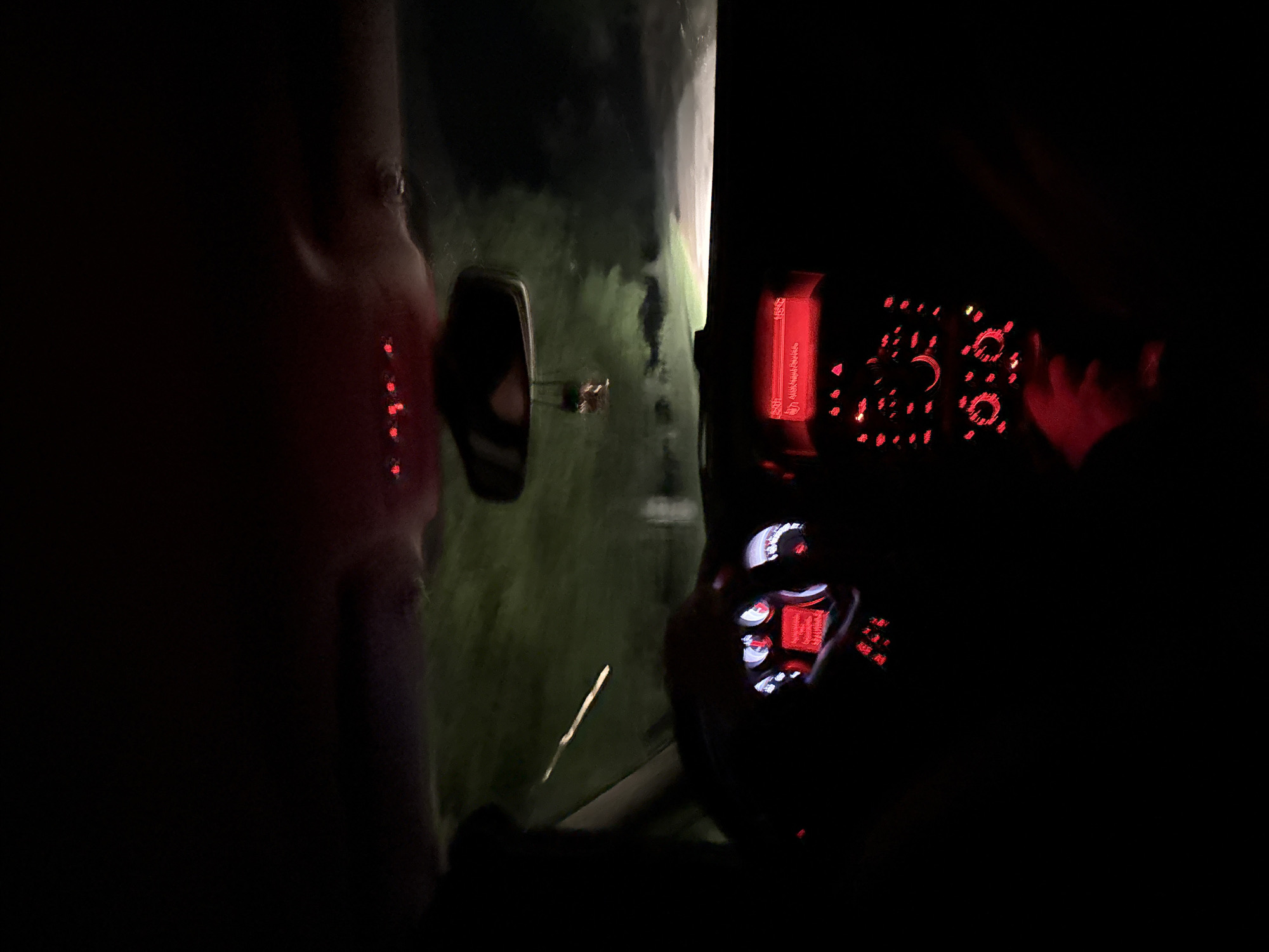 The image depicts the interior of a car driving at night in a rural area. The dashboard is illuminated with red lights. A person is visible in the driver's seat. The side mirror reflects a view of a dark road with grassy areas on the side. The location information, Slavyani, Bulgaria, is not evident in the image.