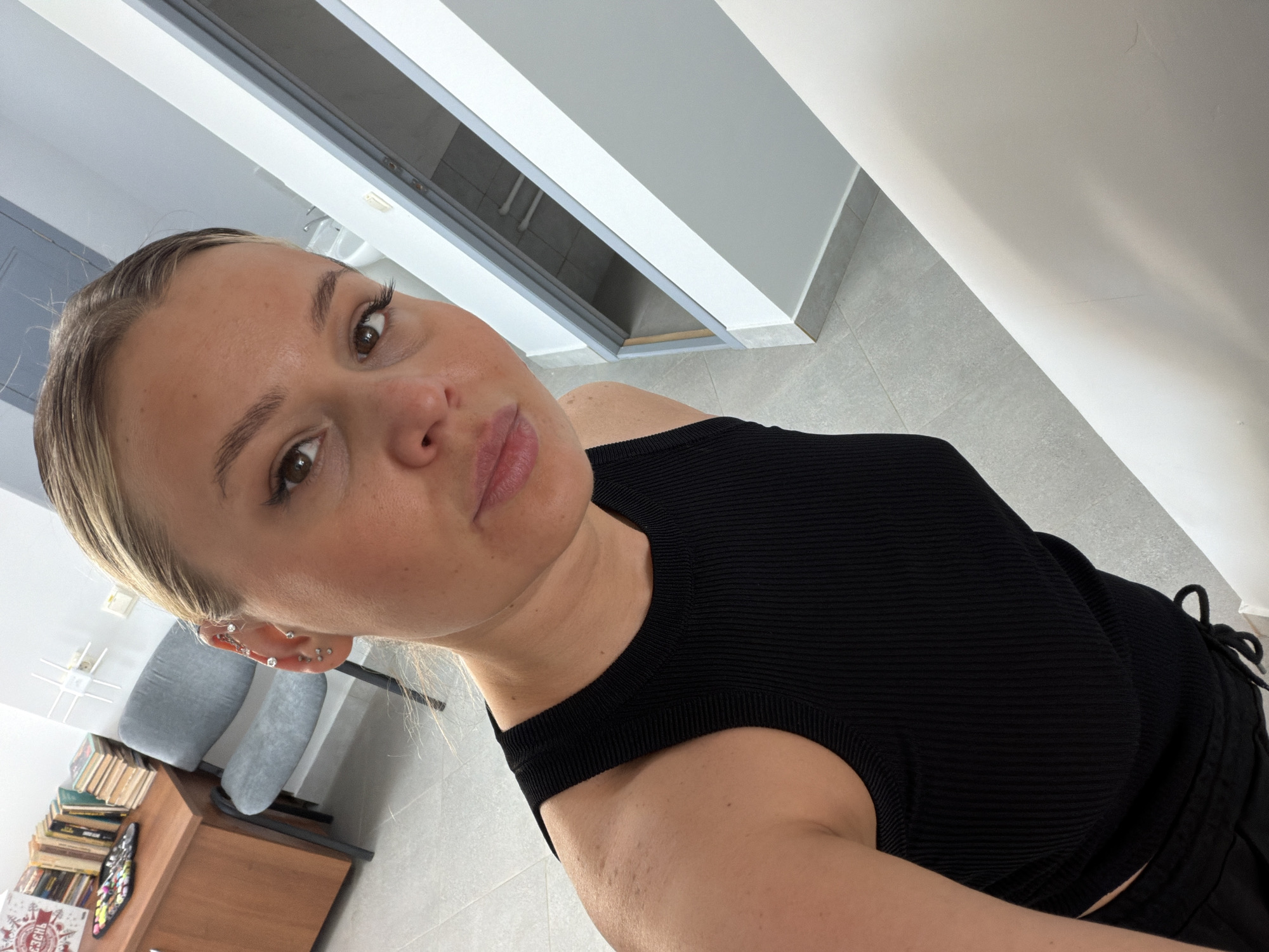A photograph taken indoors shows a fair-skinned woman with blonde hair and brown eyes looking at the camera. She is wearing a black, ribbed tank top. Her face is framed by blonde hair pulled back from her forehead. Multiple earrings are visible in her left ear.
In the background, there is a wall with white and gray horizontal stripes, suggesting modern interior design. A glimpse of a tiled floor is also visible. To the left, there's a chair, a cabinet, and a stack of books.
The image does not contain sufficient information to determine a specific location in Sarapul, Russia.