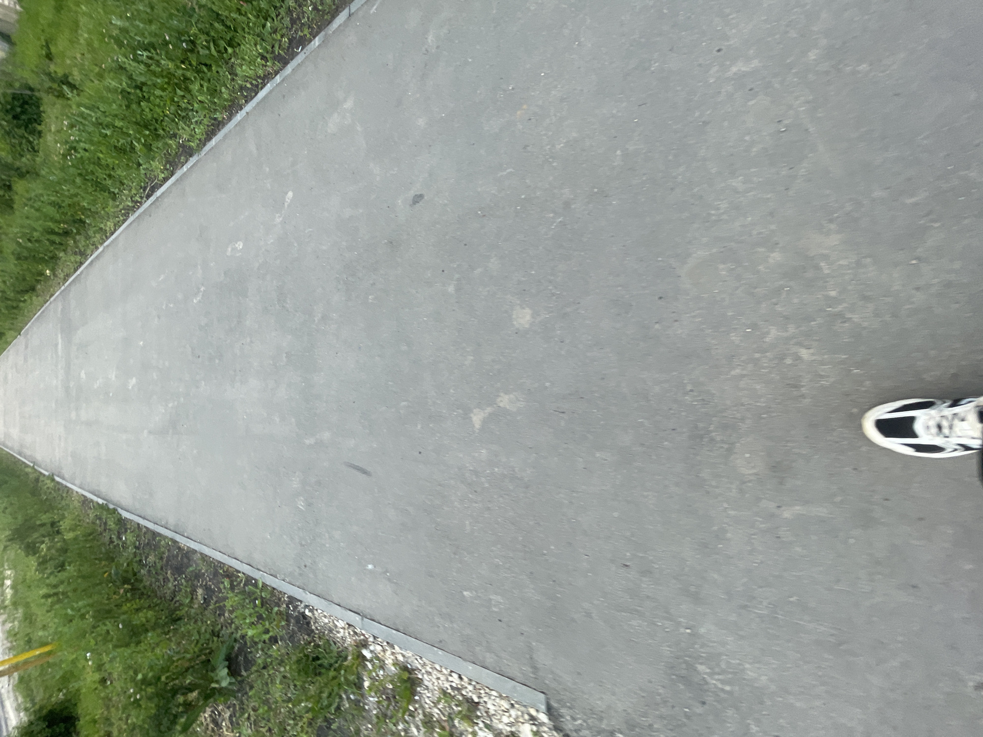 The image shows a first-person perspective of someone walking on an asphalt path, likely a bike or pedestrian lane, in Красные Баки, Russia. The path is bordered by grass and greenery on either side. In the foreground, a black and white athletic shoe is visible, indicating the action of walking. The path extends into the distance, suggesting an outdoor recreational area or a path within the town. The lighting suggests it is daytime.