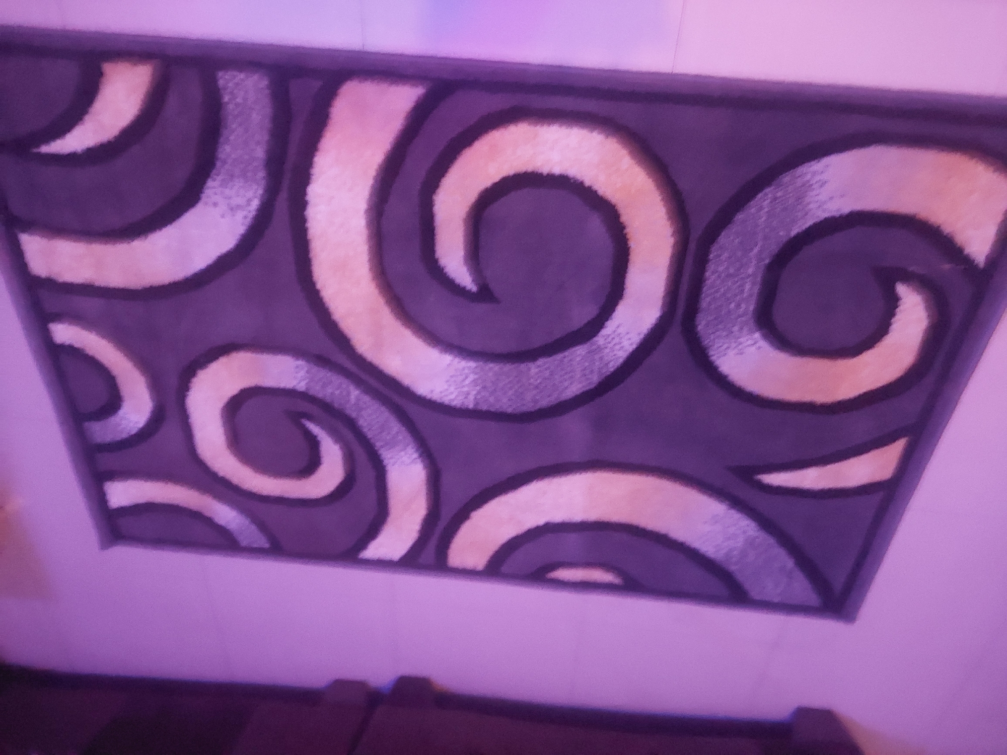 An overhead shot displays a dark rug with a swirling pattern. The rug features bold, dark outlines that define large, abstract spiral shapes. Within these spirals, there are alternating bands of light beige and a textured, lighter gray, creating a sense of depth and movement. The rug is positioned on a light-colored tiled floor. To the left of the rug, a dark wooden furniture piece is partially visible. The overall lighting in the scene casts a purple hue over the image. The photograph was taken in Bukuru, Nigeria.