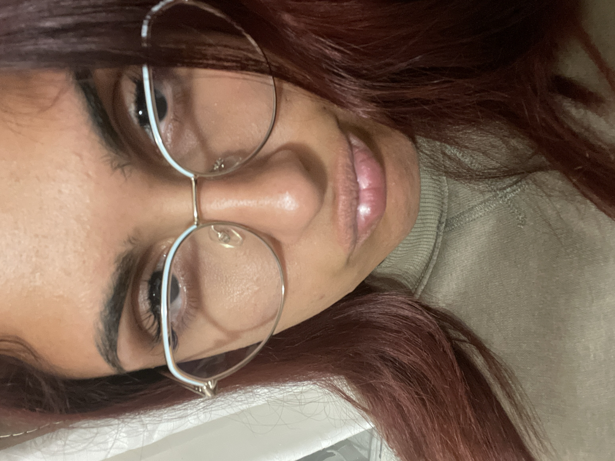 The image shows a close-up, low-angle selfie of a young woman wearing glasses. She has dark hair that appears to be dyed a reddish-brown color and is wearing a light green, ribbed turtleneck sweater. The background is indistinct but appears to be an indoor setting with some white fabric on the left, possibly a curtain. The woman's face is the primary focus, with her eyes visible through her glasses and her lips slightly pursed. There is no discernible location within the city of Varna, Bulgaria.