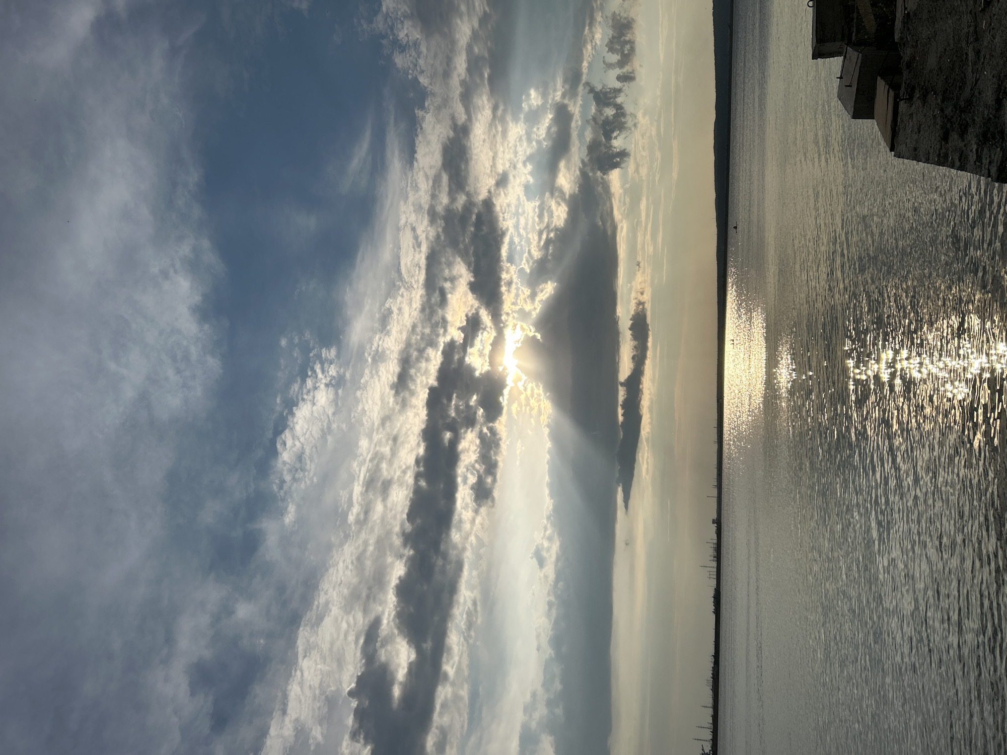 The image shows a wide shot of the Kama River on a cloudy day. The sun is partially obscured by clouds, casting rays of light across the water and creating a shimmering reflection. In the distance, the shoreline is visible with industrial structures and a silhouetted skyline. In the foreground, a concrete embankment lines the right side of the image. A lone figure is visible in a small boat on the water. The location is confirmed to be the Kama River in Chaikovsky, Russia, based on the distinctive industrial landscape visible on the horizon.