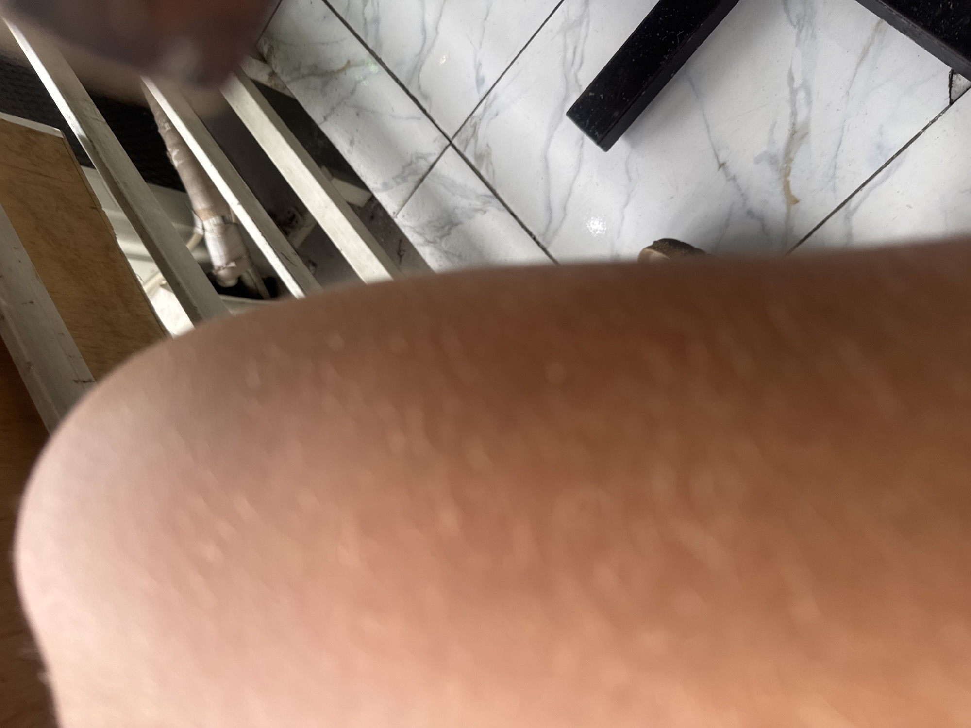 The image displays a close-up view of a person's leg, showing skin texture and some light spots. In the background, there is a white metal railing, a piece of wood, and a tiled floor with a marble pattern. A black object, possibly part of furniture, is visible on the floor. The overall scene suggests an outdoor or semi-outdoor setting, possibly a patio or a balcony. The location is identified as Kuta Selatan, Indonesia, which is a well-known tourist area.