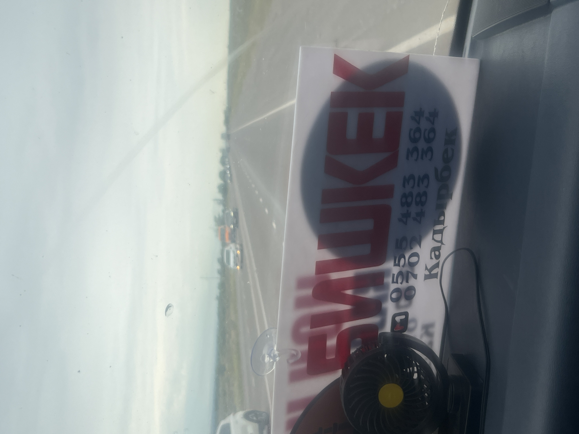 The image shows a view from inside a vehicle, looking through the windshield at a highway. The windshield is dusty and has a water spot. Attached to the windshield by a suction cup is a white sign with large red lettering and some numbers and text in black. The red lettering appears to be part of a word, with the visible portion reading "БИШКЕК". Below this, there are two phone numbers: "0555 483 364" and "0702 483 364". Beneath the phone numbers, in black text, is the word "Кадырбек". A small black electric fan with a yellow center is visible in the lower left corner of the image, resting on the dashboard. The background shows a highway with other vehicles in motion under a cloudy sky. The landscape appears to be flat and rural. The presence of "БИШКЕК" (Bishkek) on the sign indicates a likely connection to the capital city of Kyrgyzstan, suggesting the image may have been taken on a route leading to or from Bishkek, or within its vicinity.