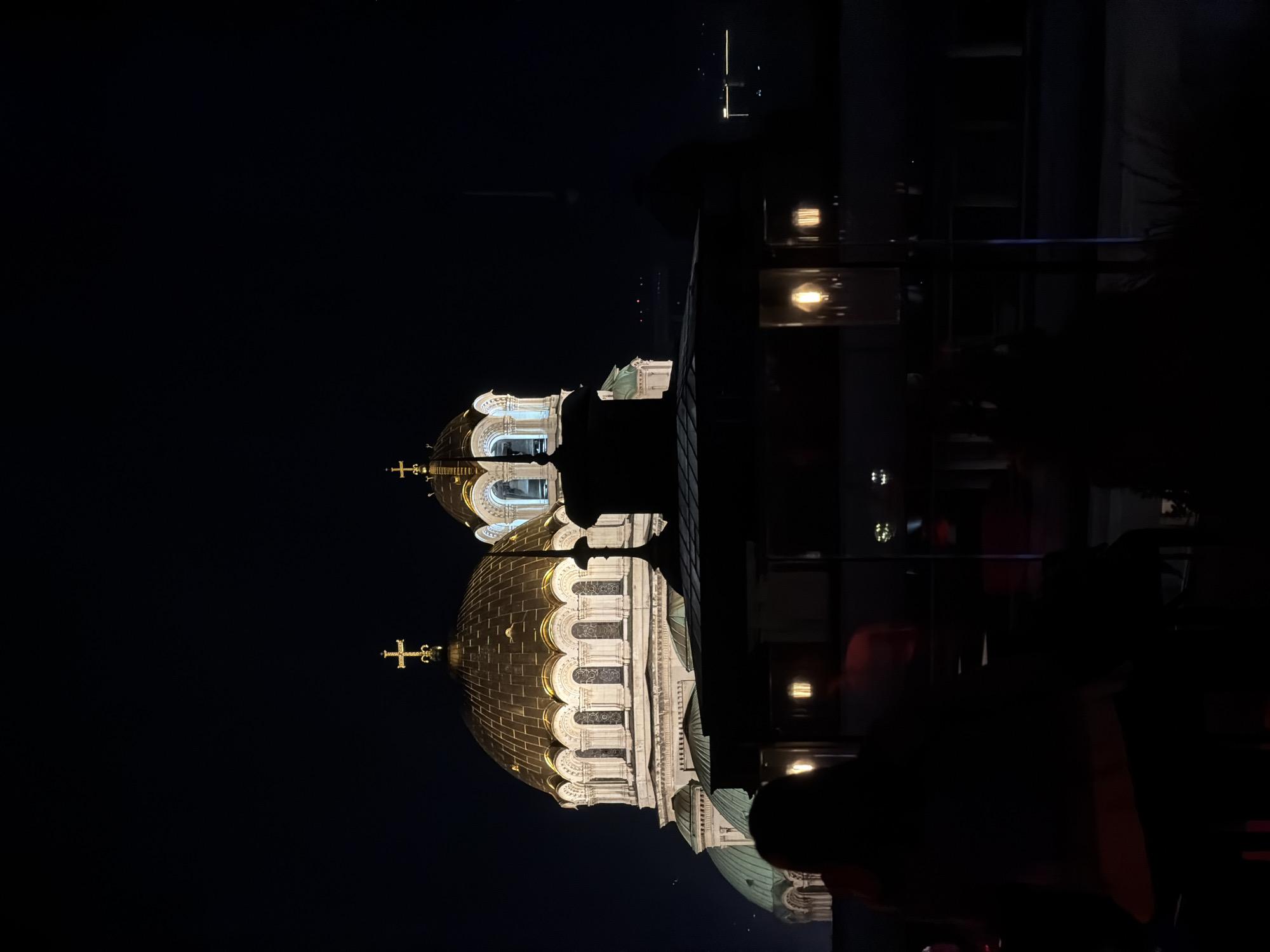 The image depicts the illuminated domes of the St. Alexander Nevsky Cathedral in Sofia, Bulgaria, at night. The domes, topped with golden crosses, are the primary focus, reflecting light against the dark sky. The reflection of the dome on the glass shows the dark green color of the roof. The image seems to be taken from a higher vantage point, possibly a building, looking down at the cathedral. Silhouetted figures are visible in the foreground, suggesting the presence of people observing the scene. The background is dark.