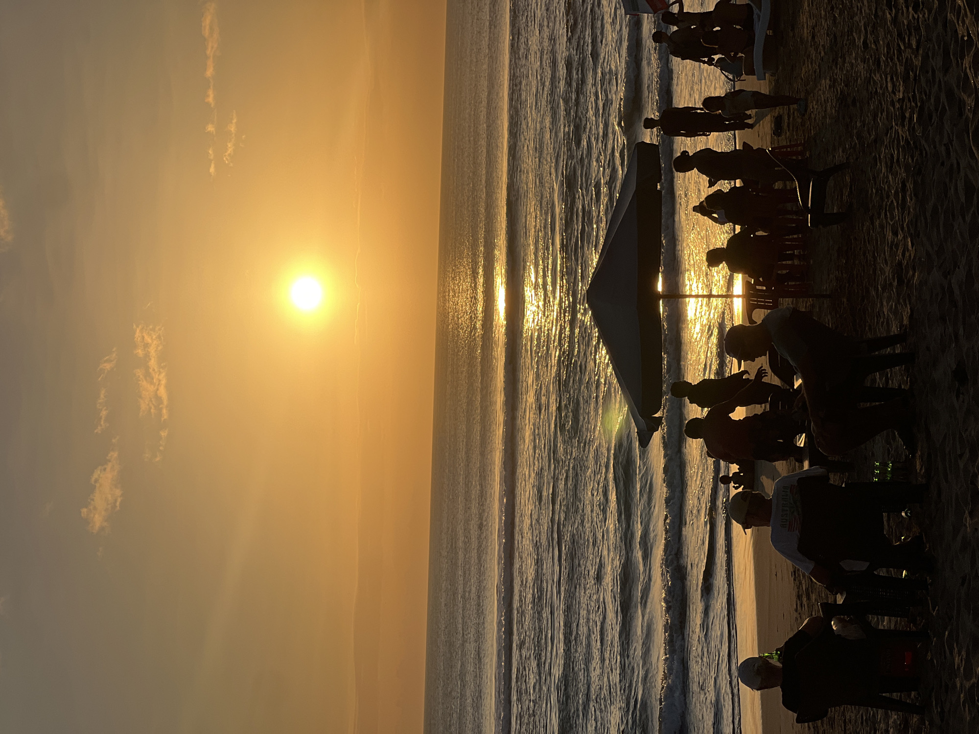 The image depicts a sunset on Kuta Beach, Bali, Indonesia. Silhouetted figures of people are scattered across the sandy beach, some sitting in chairs, others standing. A large umbrella provides shade for a group gathered at tables. Gentle waves roll in from the ocean, reflecting the warm, golden light of the setting sun. The sky is a gradient of orange and yellow, with a few wispy clouds. The overall atmosphere is serene and tranquil, characteristic of a beach sunset.