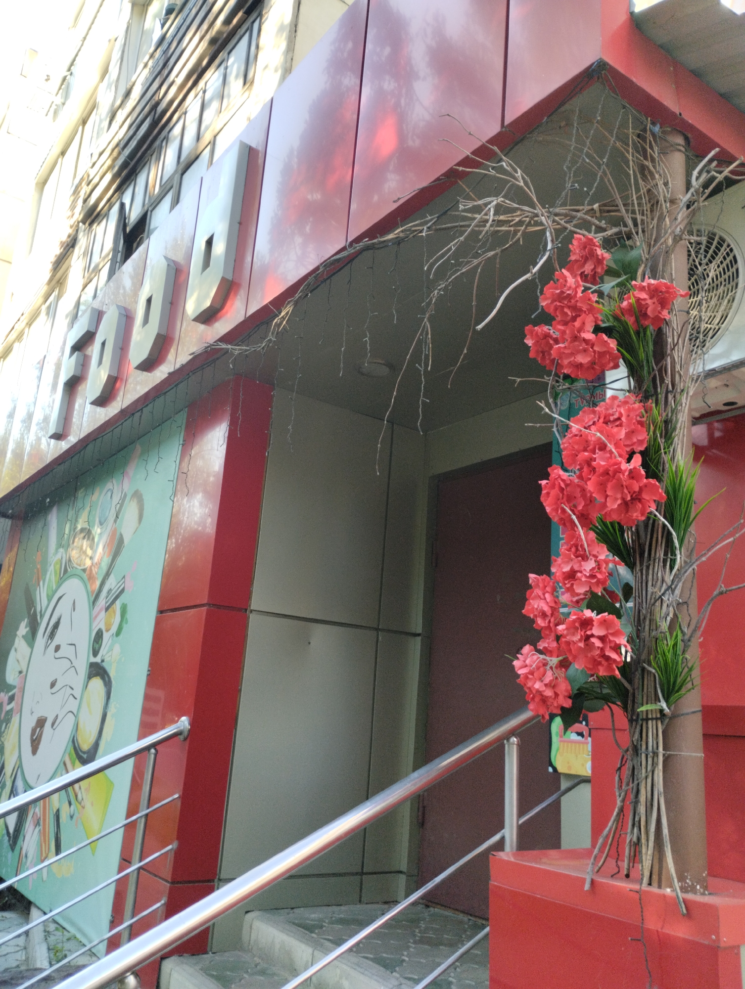 The image shows the exterior of a building with large, raised white letters spelling "FOOD" on a red facade. Below the sign, there is a colorful mural or advertisement featuring what appears to be a stylized face with makeup items like brushes and lipstick. A metal railing with multiple bars leads up a set of stairs, flanking the entrance. To the right of the entrance, a decorative arrangement of dried branches and clusters of red artificial flowers is attached to a pole. The building appears to be in an urban setting, with a light-colored wall and windows visible on the upper floors. The overall impression is of a commercial establishment, possibly a restaurant or food-related business, in Kostanay, Kazakhstan.