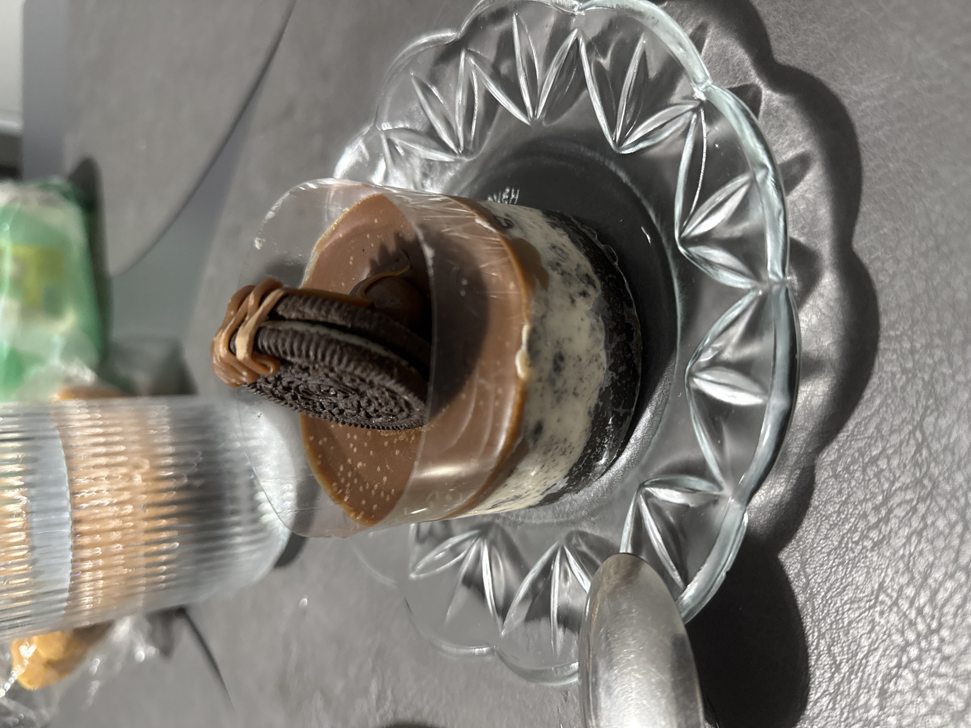 The image depicts a dessert, likely a layered cake or mousse, on a clear, ornate glass plate. The dessert appears to be comprised of dark cookie layers and a light, creamy layer, topped with a brown, glossy topping and two Oreo cookies. The dessert is partially enclosed in a clear plastic wrapper. To the left of the dessert, out of focus, is a stack of what appear to be wrapped cookies or wafers. The background is a gray, textured surface, possibly a table or counter. The image doesn't provide any clues to suggest its location is Stara Zagora, Bulgaria. The description is purely based on the visible elements in the photo.