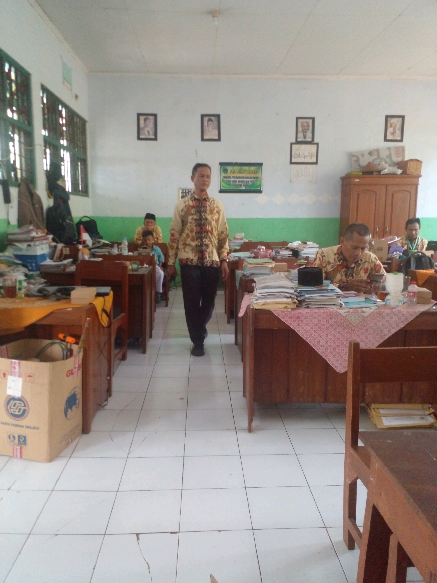 The image depicts the interior of what appears to be a classroom in Banyuputih, Indonesia. A man in a patterned shirt and dark pants is walking towards the camera. Several students and other individuals are seated at desks, some of whom are looking at the camera. Desks are arranged in rows, laden with books, papers, and personal belongings. A cardboard box is visible in the foreground. The walls are painted white and green, adorned with framed pictures and a green sign. The floor is tiled and appears to have some cracks.