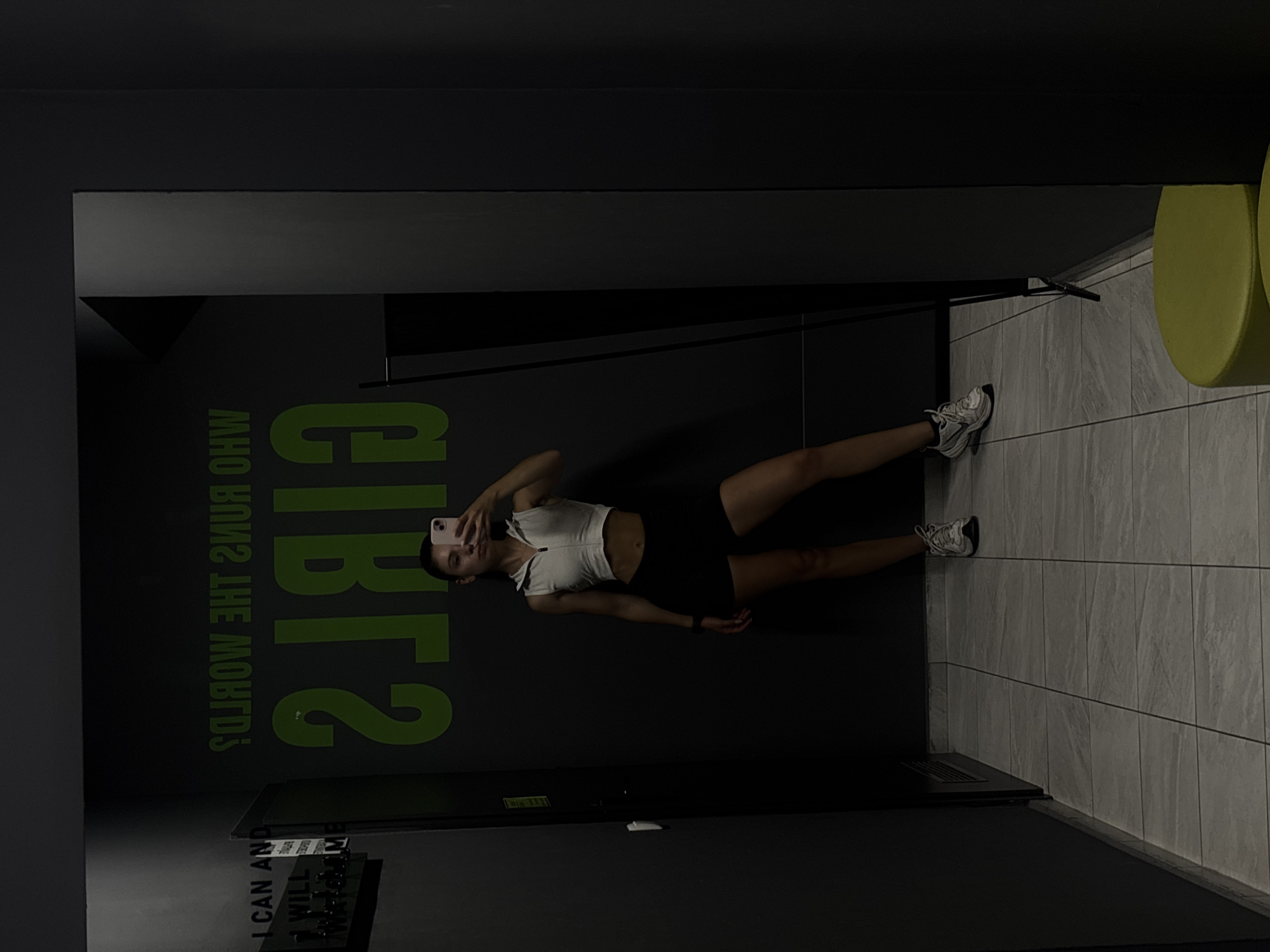 A young woman is captured taking a selfie in a gym locker room. She is wearing a white crop top with a zipper and black athletic shorts. The background features a dark gray wall with large, bright green lettering that reads "GIRLS RUN THE WORLD" in reverse. To the left of the woman, also on the wall, is text that reads "I CAN AND I WILL Watch ME." The floor is tiled with a gray, stone-like pattern. She is standing in front of a mirror, her reflection visible. The lighting is dim, creating a moody atmosphere. The scene appears to be inside a fitness facility in Varna, Bulgaria.