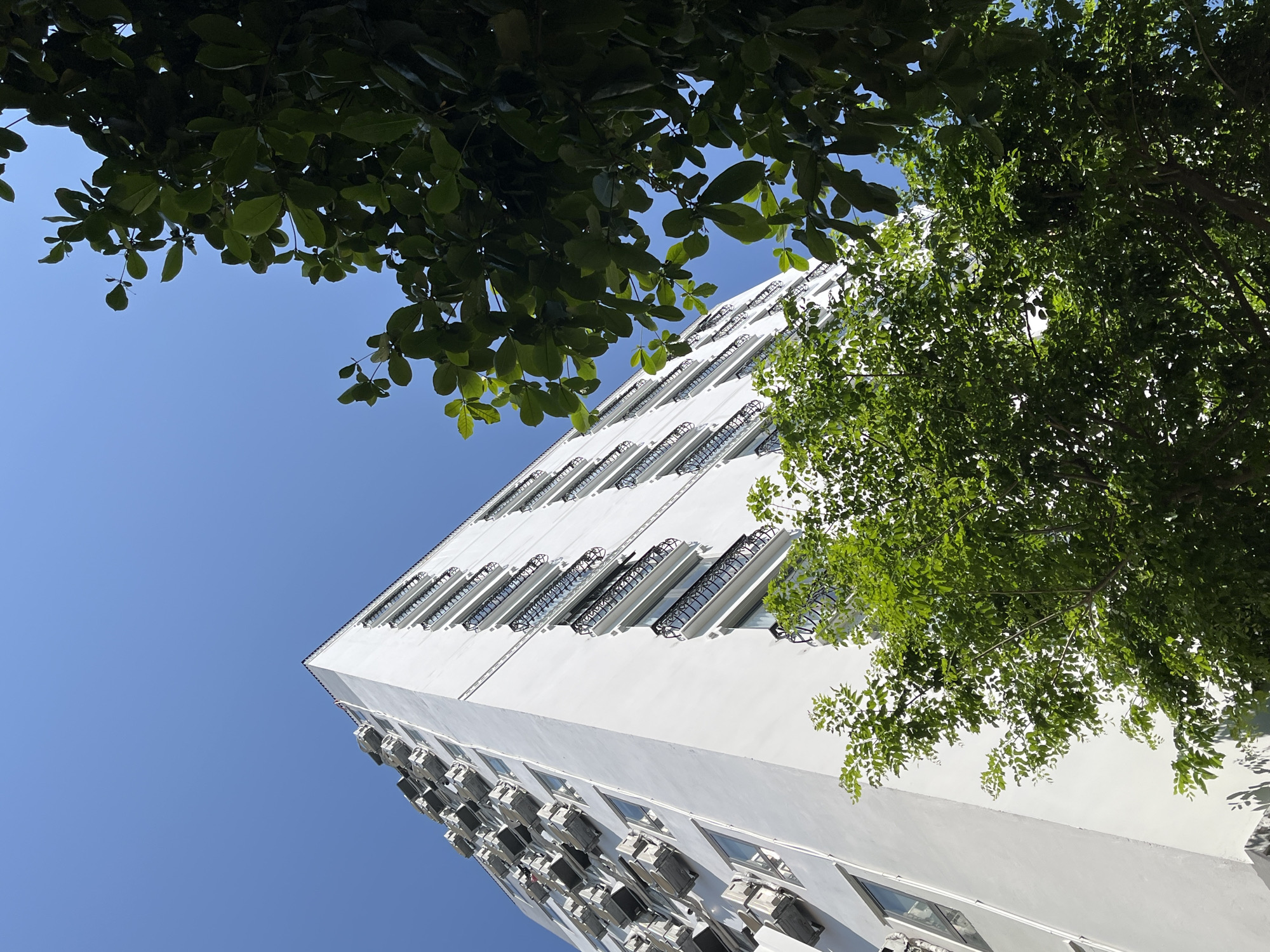 A low-angle view depicts a tall, white building under a clear, blue sky. The building has numerous windows and small balconies with black railings. Many air conditioning units are visible on the lower portion of the building. Leaves from trees frame the top of the image. The photo was taken in Đà Nẵng, Vietnam.