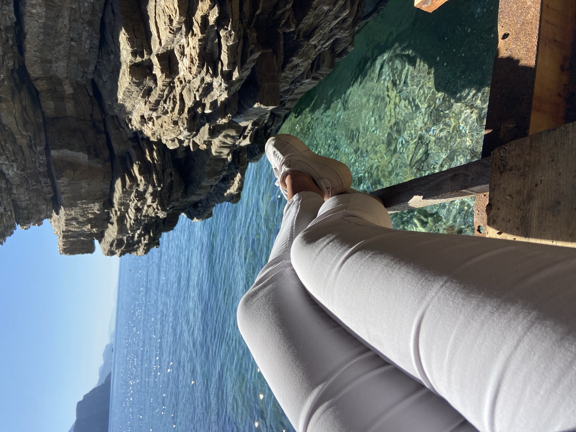 A person wearing white pants and white Nike sneakers sits with their legs crossed on a wooden structure, possibly a small dock or platform. The location appears to be near or above clear water, likely part of the Adriatic Sea. A rocky cliff or cave formation is visible to the left, providing shade. The water is turquoise with visible textures. The location is in Budva, Montenegro.