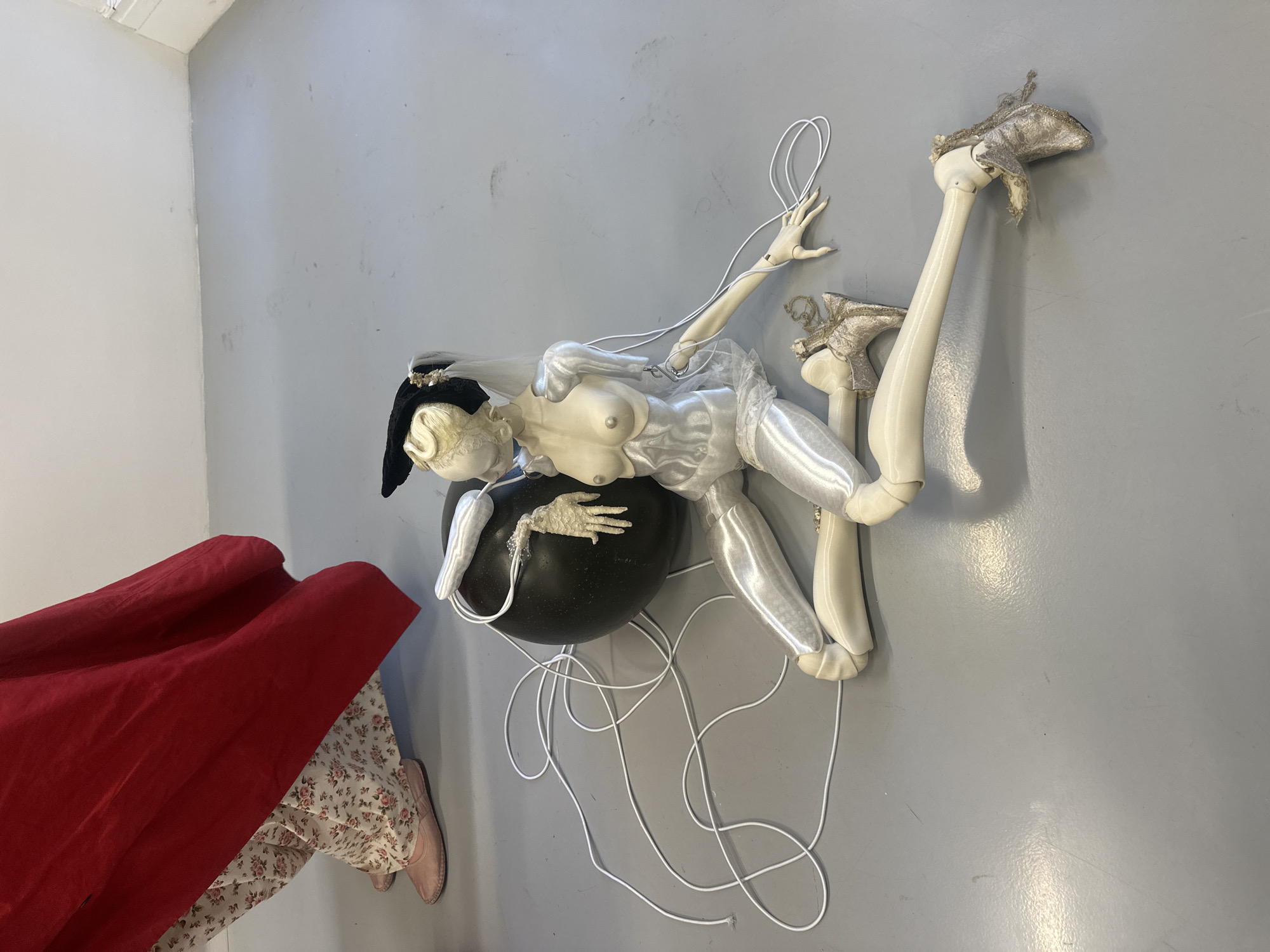 This media file captures an artistic installation or sculpture displayed in an indoor setting, characterized by plain grey walls and flooring, possibly an art studio or gallery space.
The primary subject is a stylized, off-white, mannequin-like figure with articulated limbs, resembling a large doll or puppet. The figure has blonde, wavy hair and wears a black hat or headpiece. Its upper torso is bare, featuring prominent, stylized breasts, while the lower body is dressed in shiny, white satin-like fabric and translucent ruffles. The figure is positioned in a reclining and somewhat contorted pose, with its upper body resting on a large, smooth, dark spherical object. White cords or strings are loosely draped around its limbs and scattered on the floor, suggesting connections akin to a marionette. Its feet are adorned with ornate, high-heeled shoes or boots covered in a textured, possibly beaded or sequined, fabric.
In the bottom left of the frame, the legs of a person are partially visible, wearing light-colored pants with a floral pattern and soft pink flat shoes. A large, flowing piece of rich red fabric, possibly a cloak or drape, covers part of this person and extends into the foreground, providing a strong color contrast.
The scene appears to be a static display, possibly part of an exhibition or creative process. The lighting is bright and even, typical of an interior space. No specific time of day or weather is indicated. There is no visible text in the image. The overall impression is one of a surreal and artfully arranged tableau, located in London, United Kingdom.